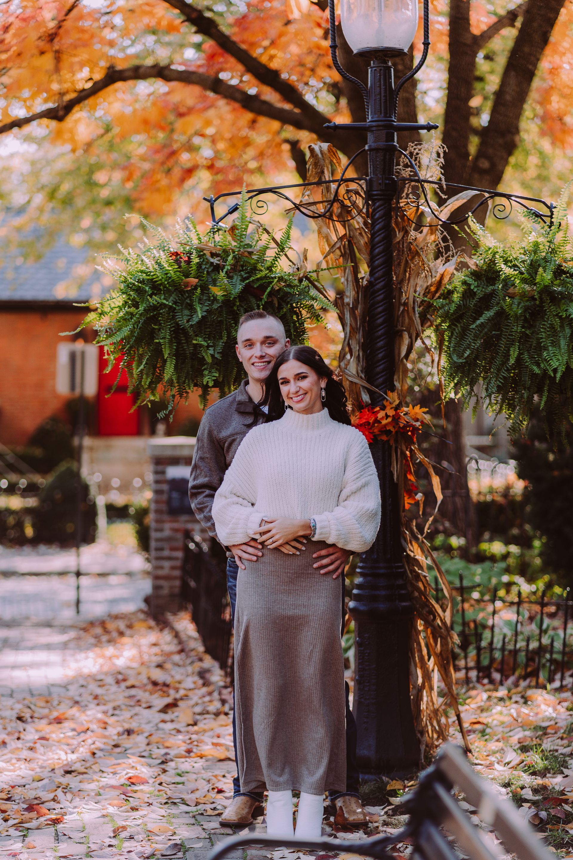 Couple in fall attire, standing by a lamppost, fall foliage backdrop.