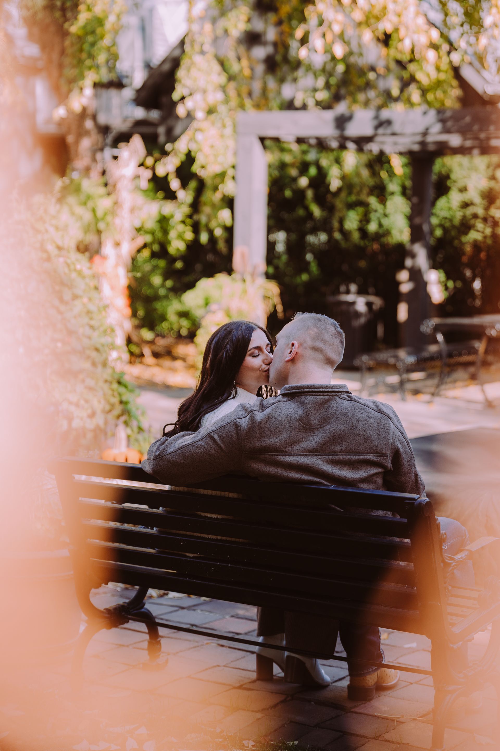 Couple kissing on a park bench. Man in a gray sweater, woman has long dark hair. Green foliage surrounds them.