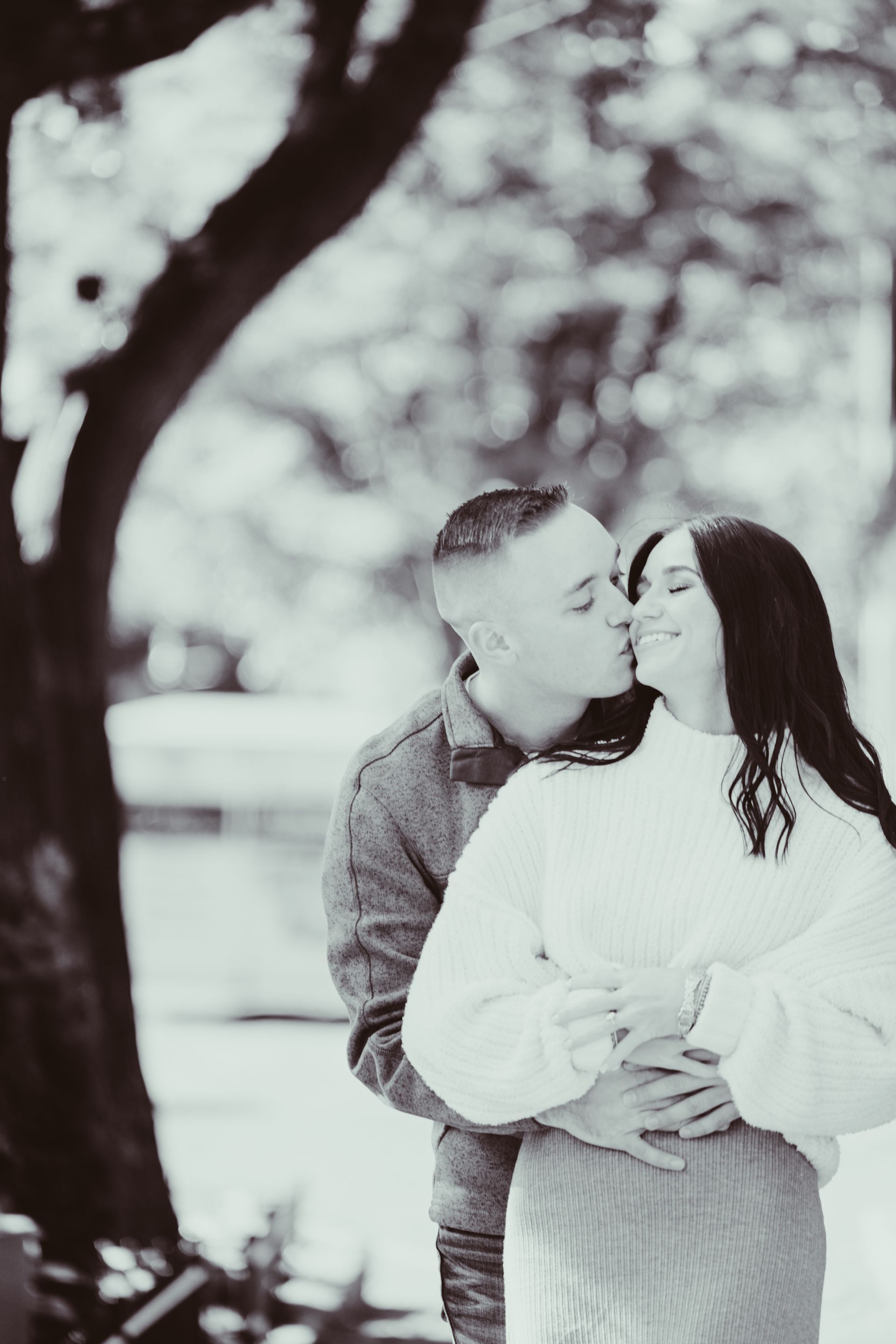 Man kissing woman's cheek; couple embracing. Outdoors by tree; black and white.