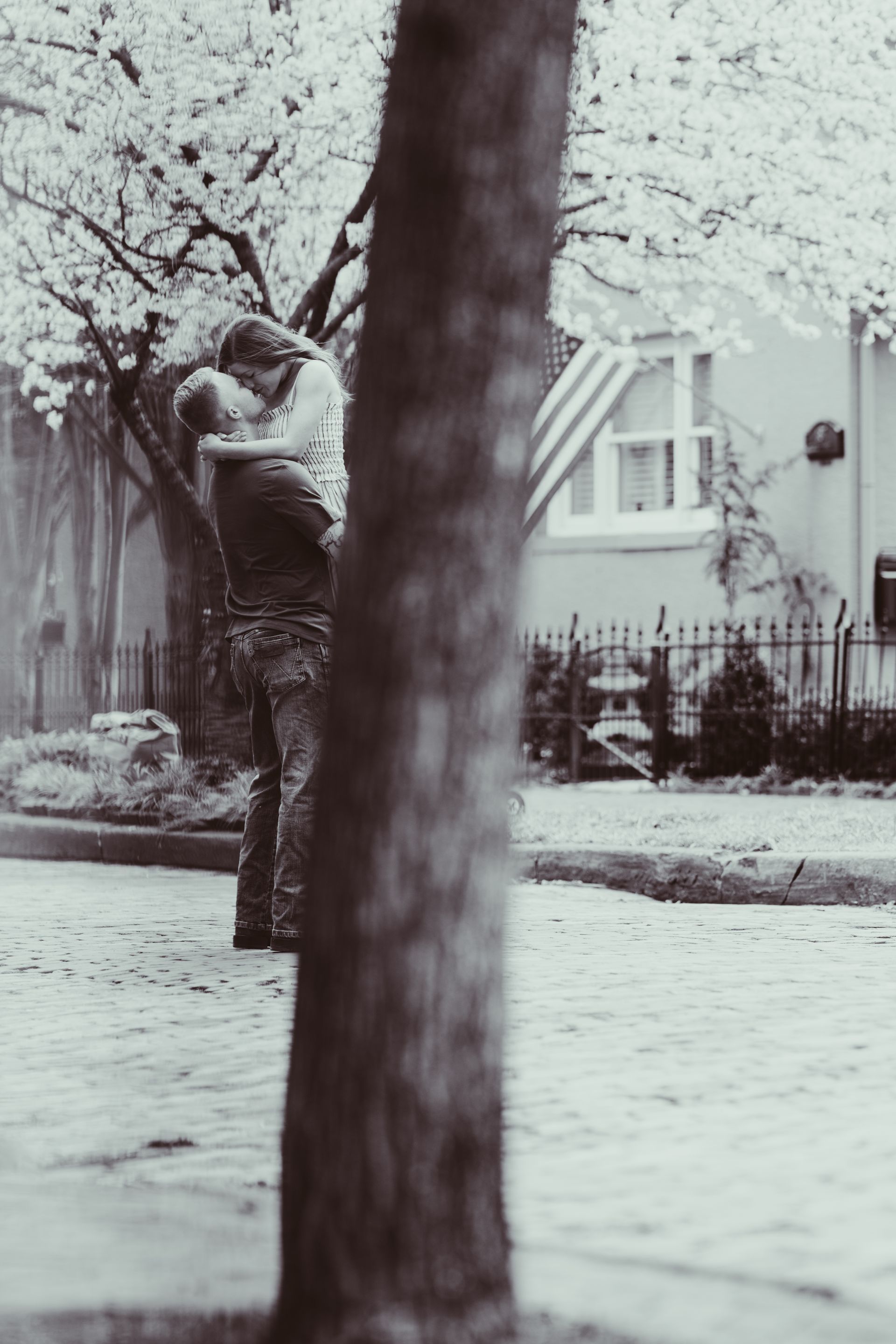 A black-and-white photo of a couple embracing and kissing outdoors, partially obscured by a large tree in the foreground.