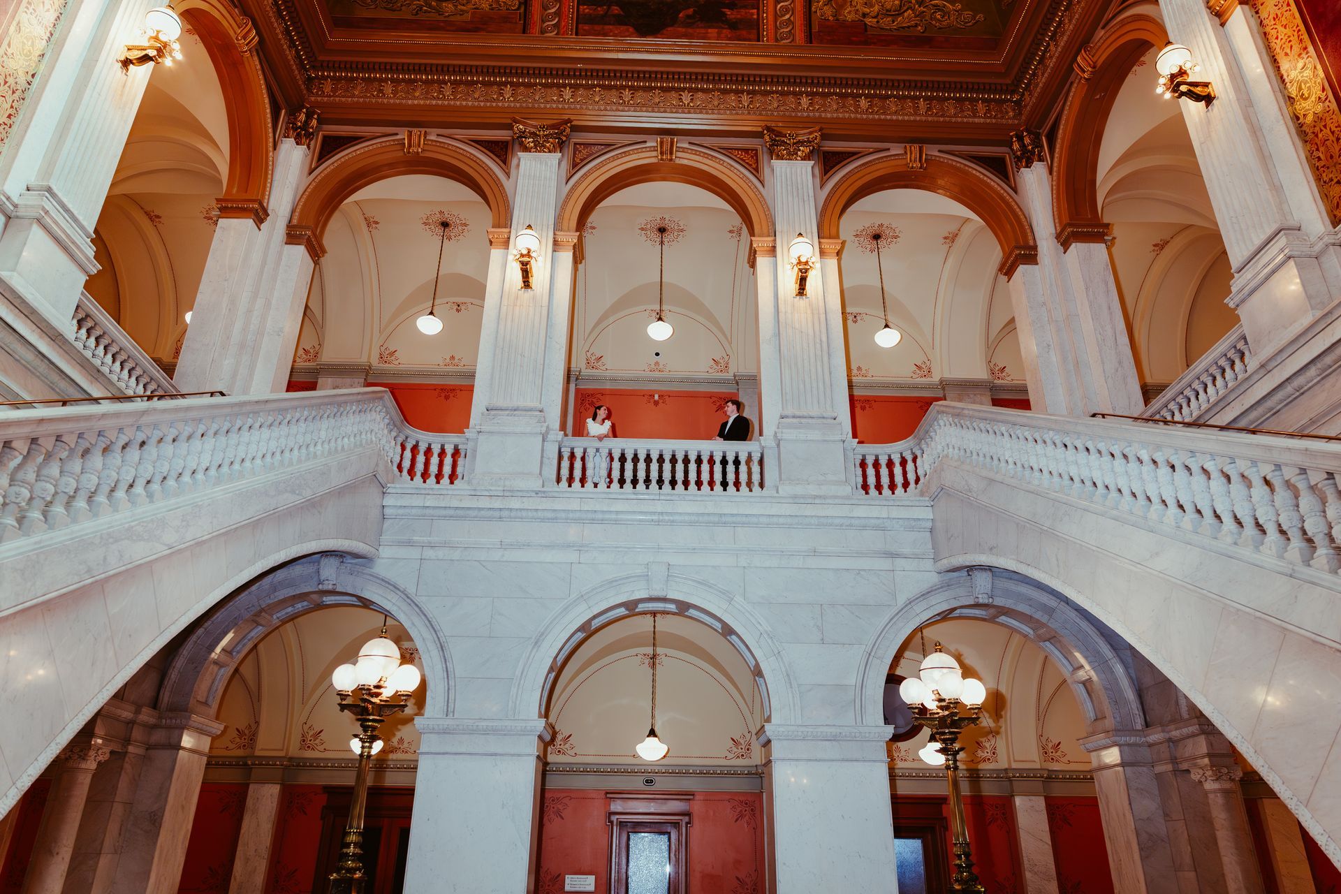 Ornate white marble staircase splits to lead up to a mezzanine with decorative arched alcoves and warm, golden lighting.