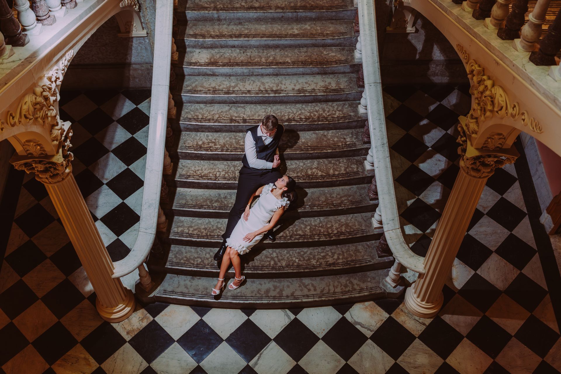 A couple sits on a grand staircase with a black-and-white checkered floor in a formal, historical setting.