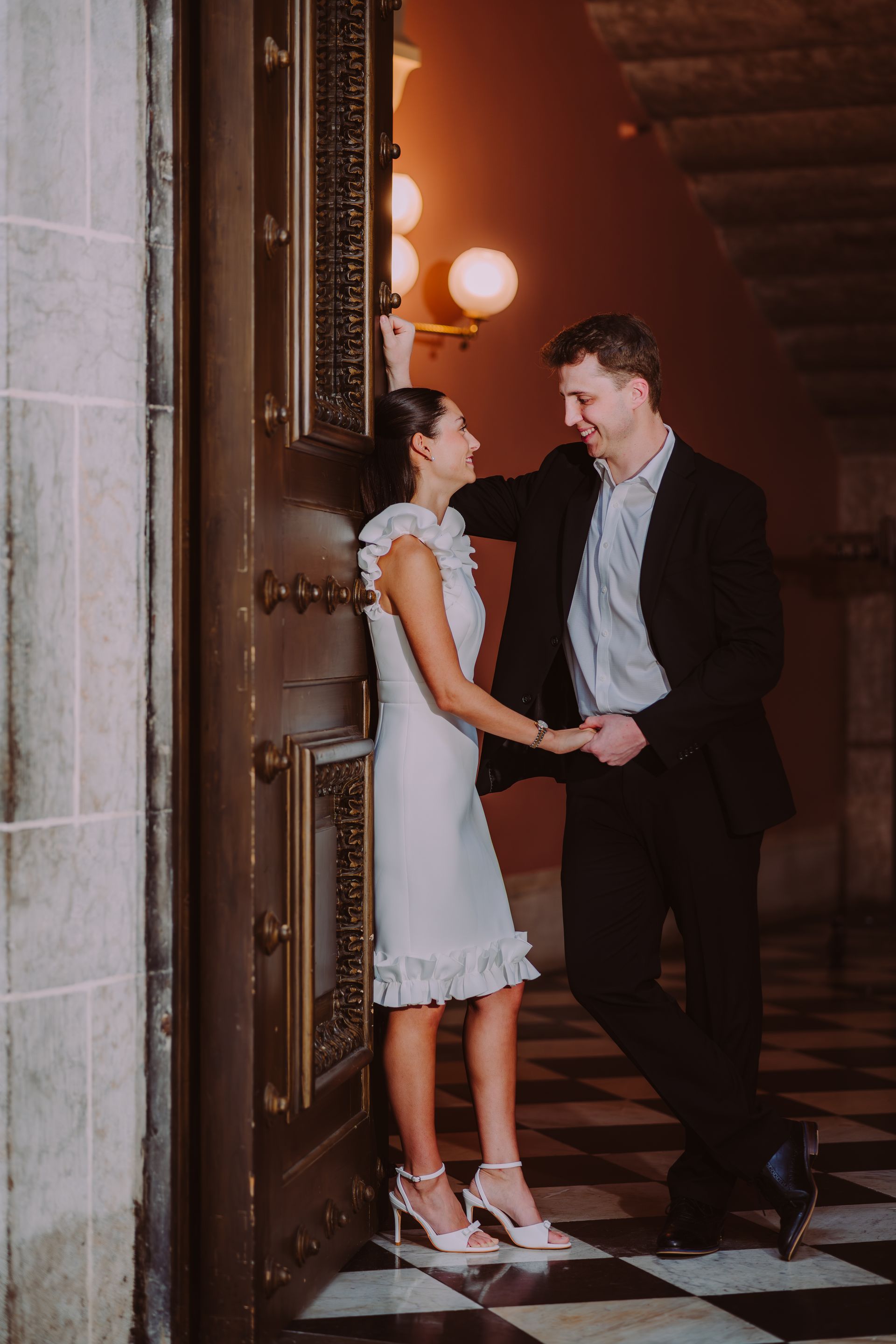 Couple holding hands in a warmly lit doorway with checkered floor and dark staircase