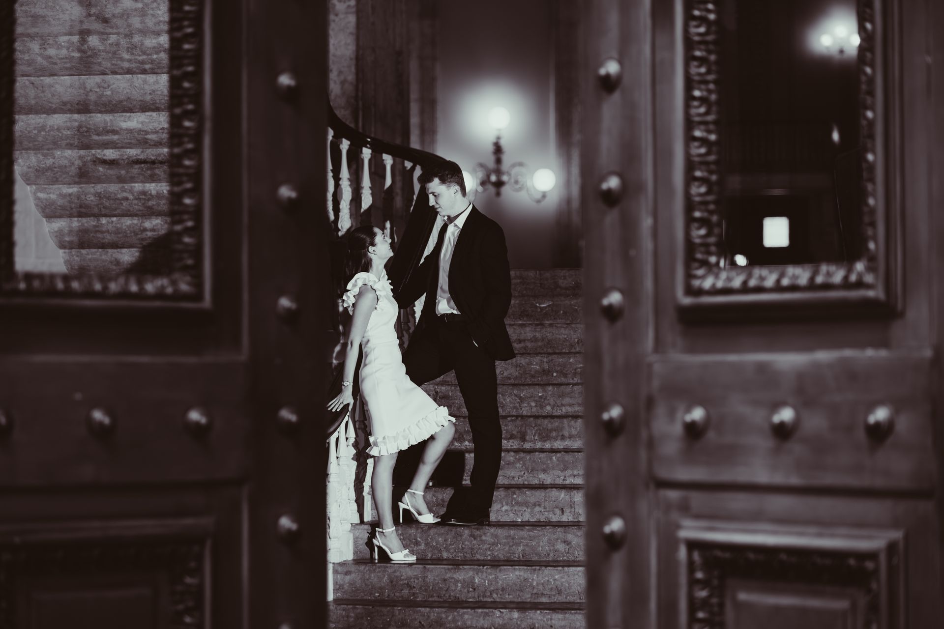 Couple descending a staircase in a dimly lit hall, seen through open wooden doors.