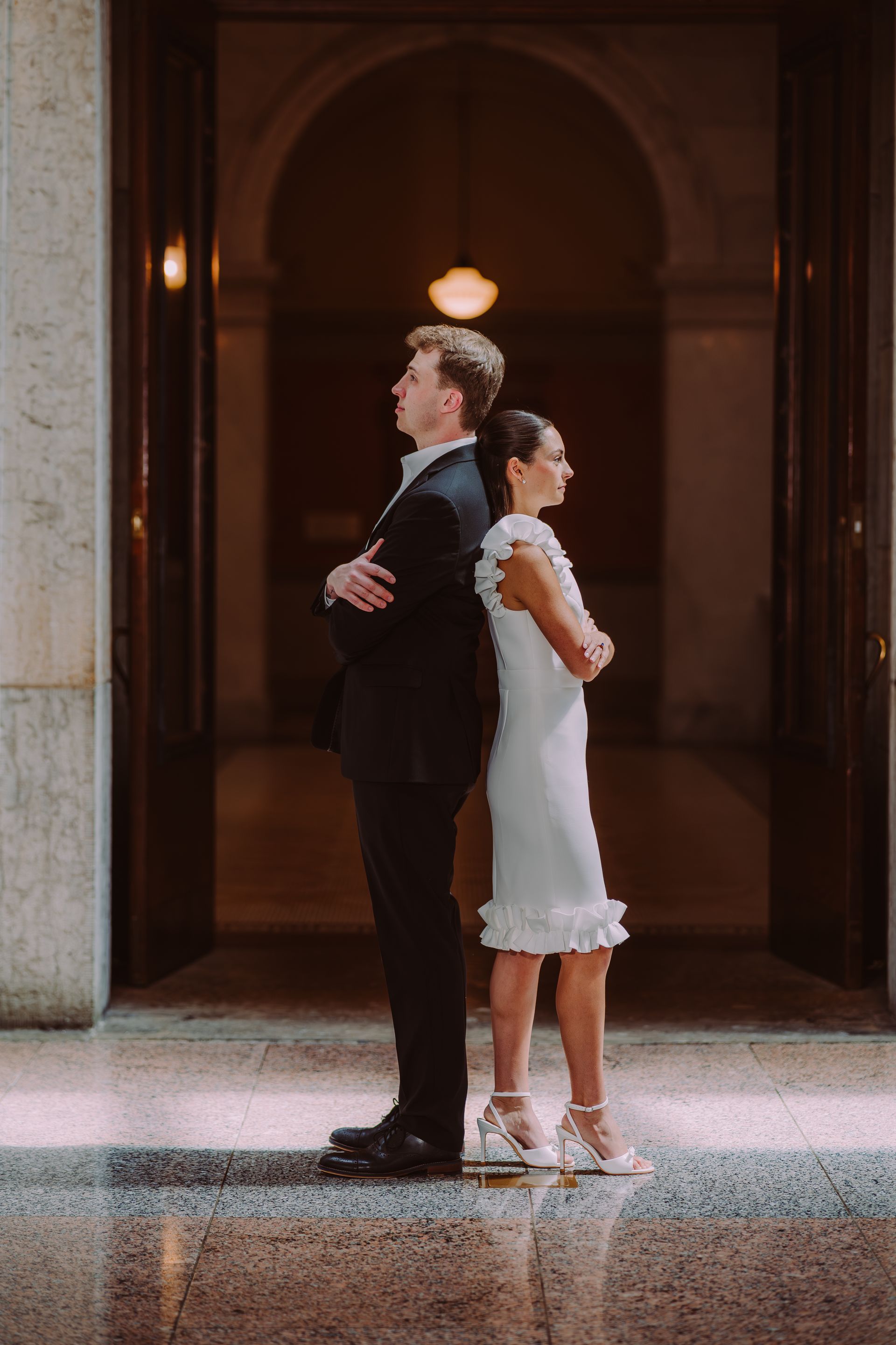 Two people in formal attire stand back-to-back in a dimly lit archway.