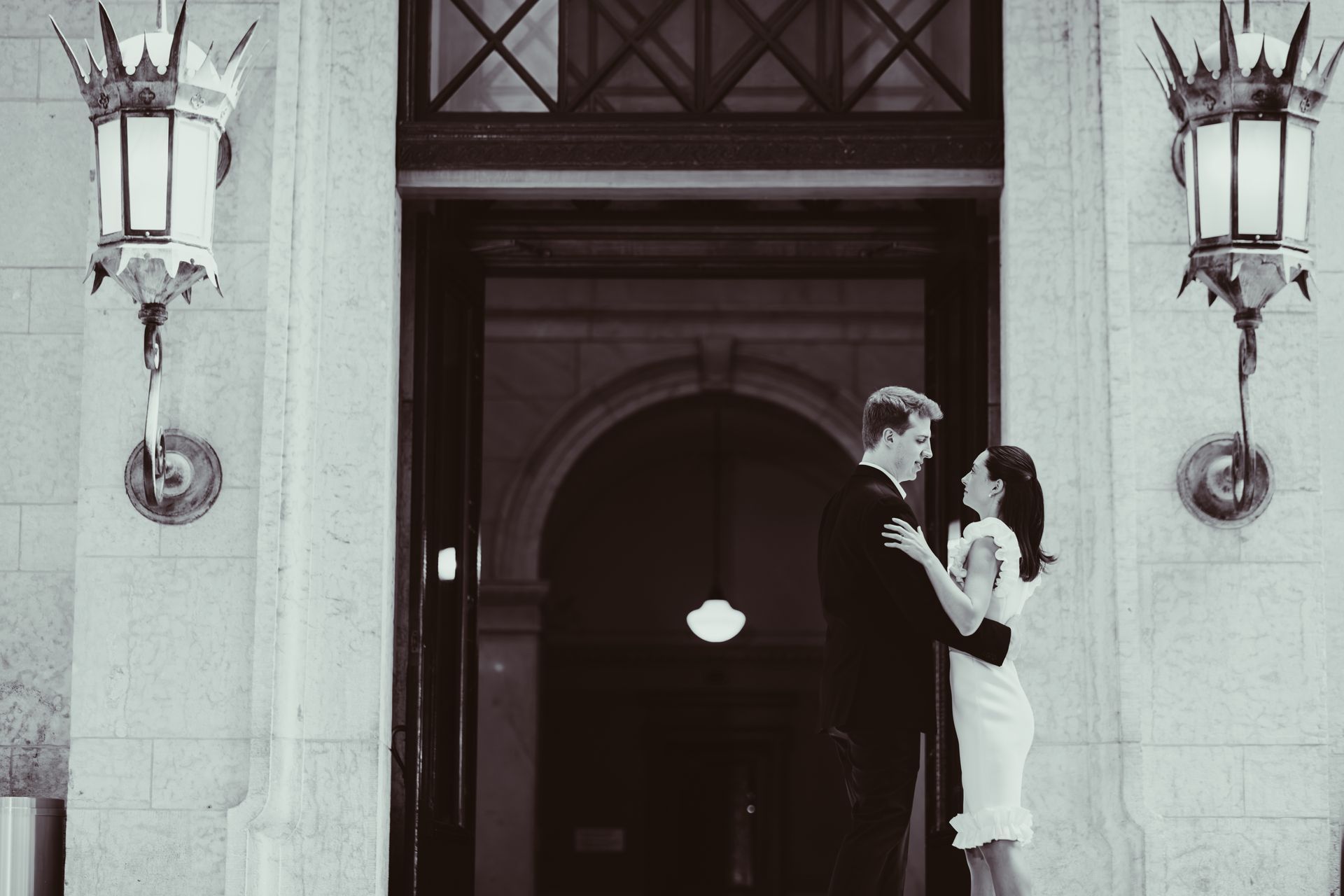 Couple embracing at a building entrance, framed by tall lanterns and an open doorway.