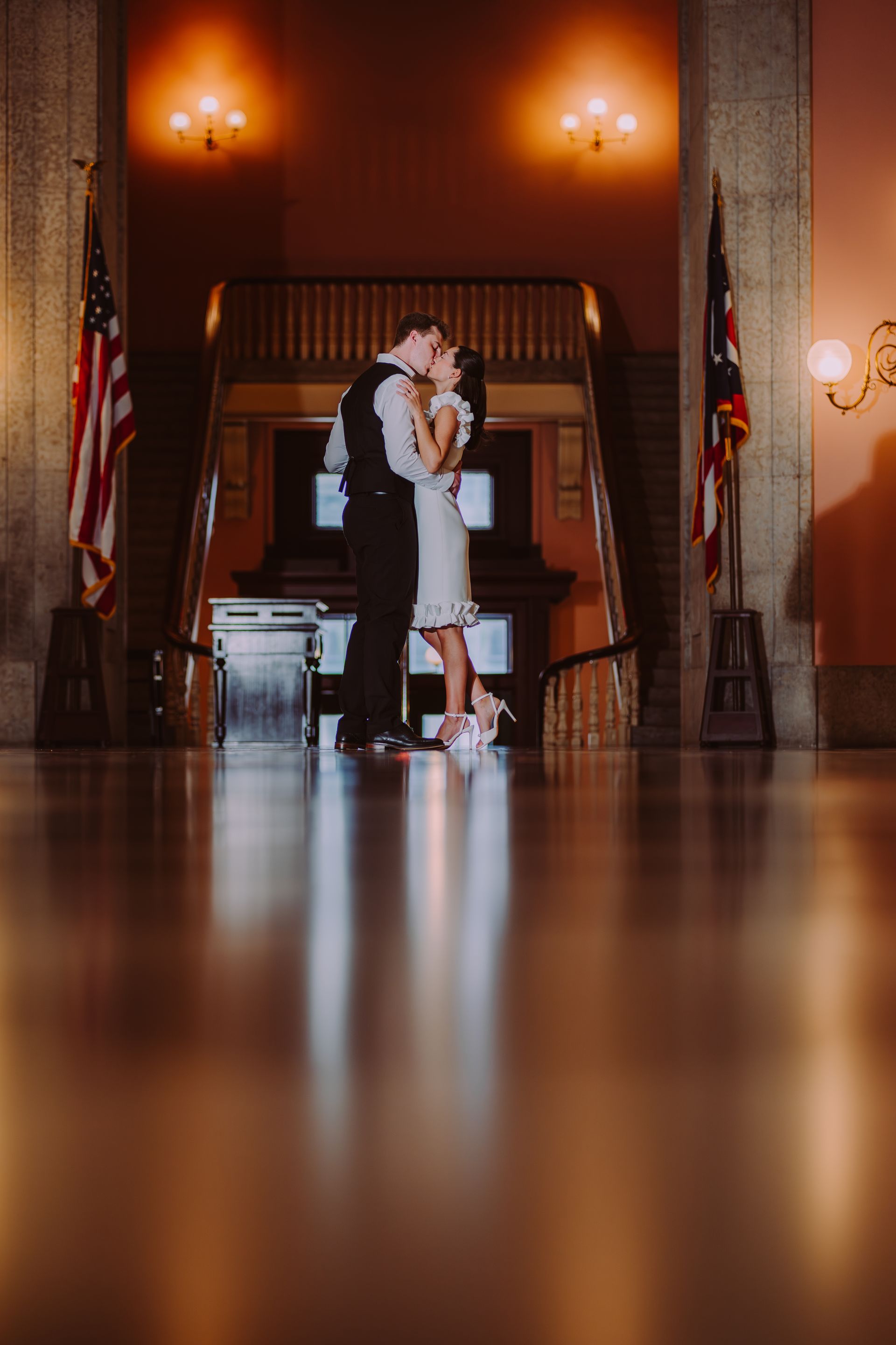 Couple embracing and kissing in a grand hall with warm wall lights and flags.