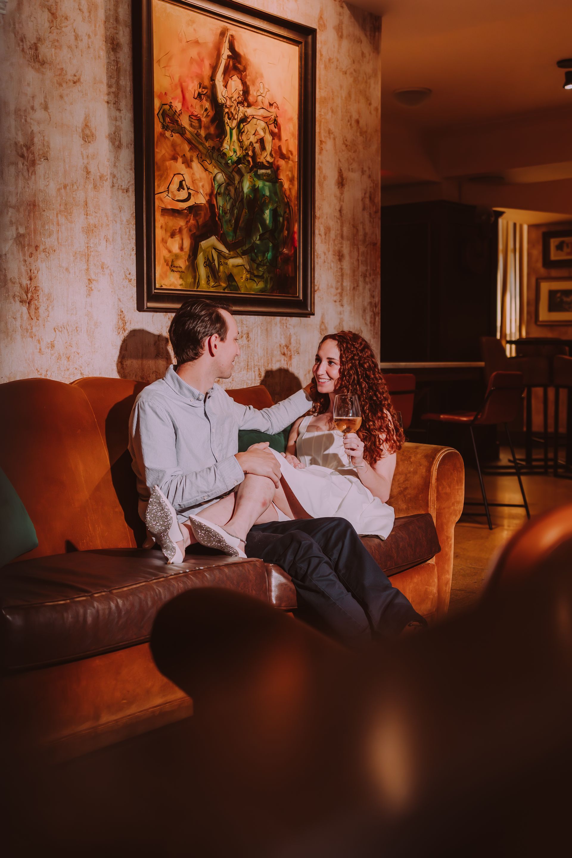 Couple on an orange couch, woman holding a glass of wine, smiling, near a painting in a dimly lit setting.
