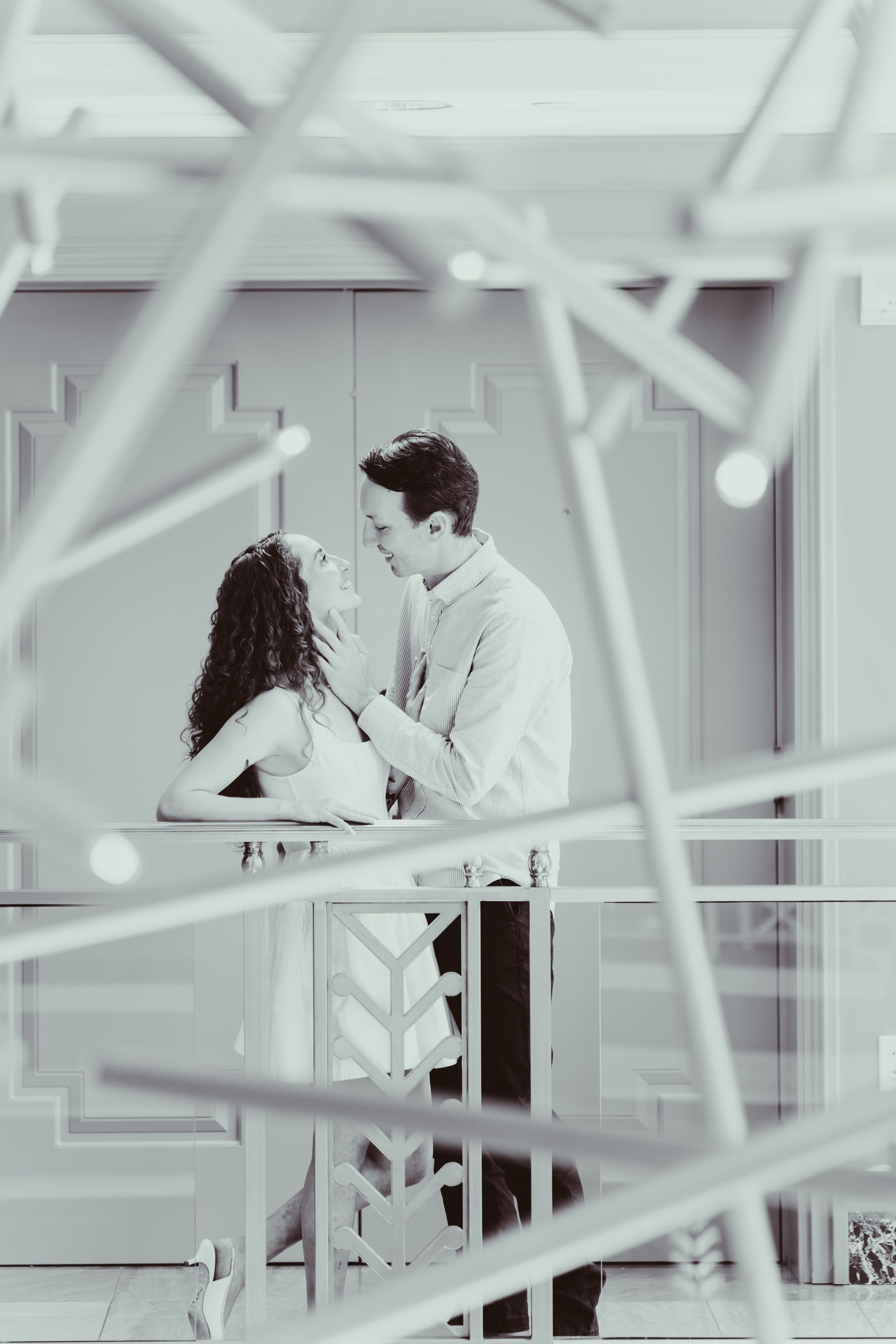 Couple gazing at each other on balcony, abstract light fixture in foreground. Black and white.