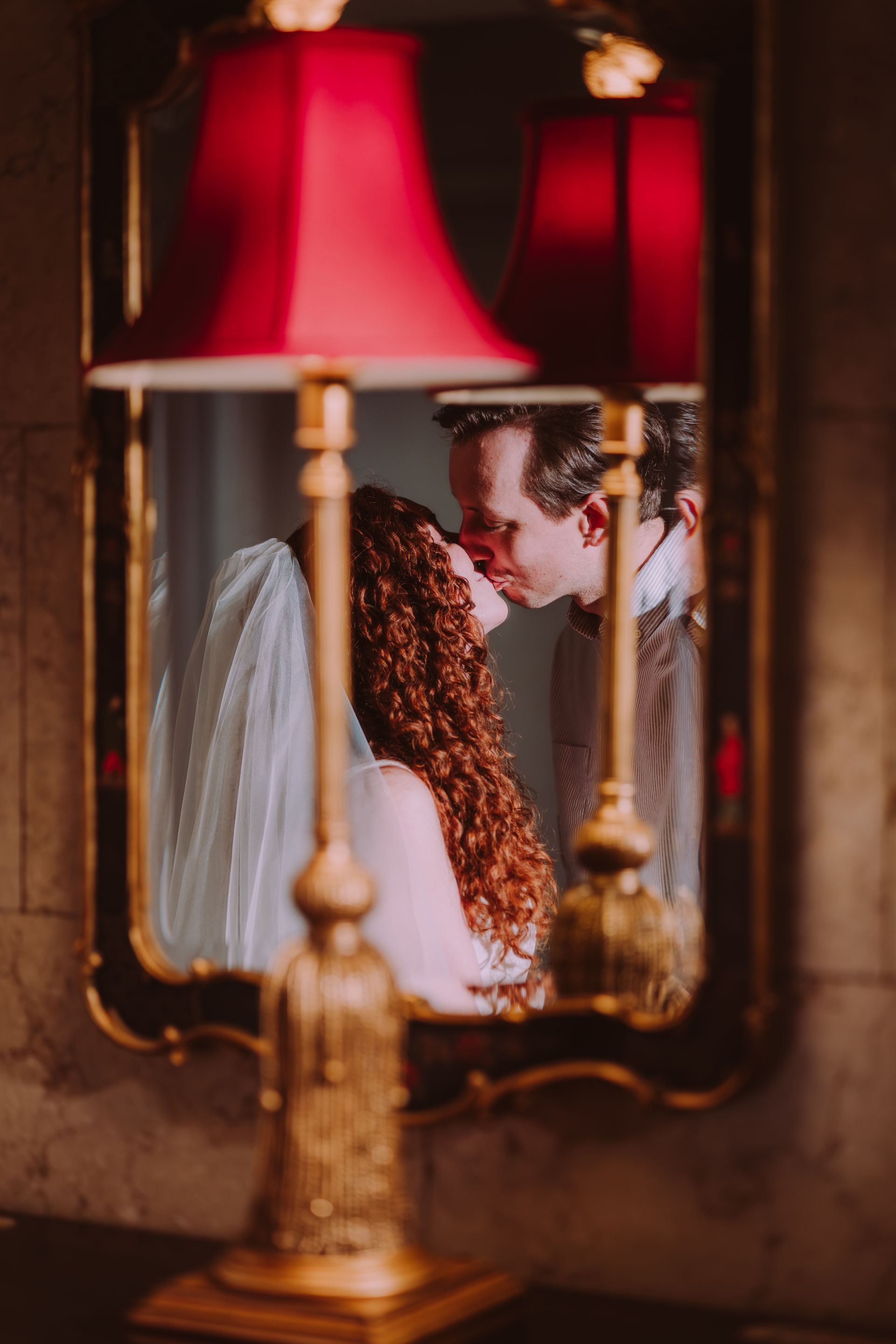 Couple kissing in ornate mirror, framed by red lamps. Bride with curly hair, groom with suit, intimate moment.