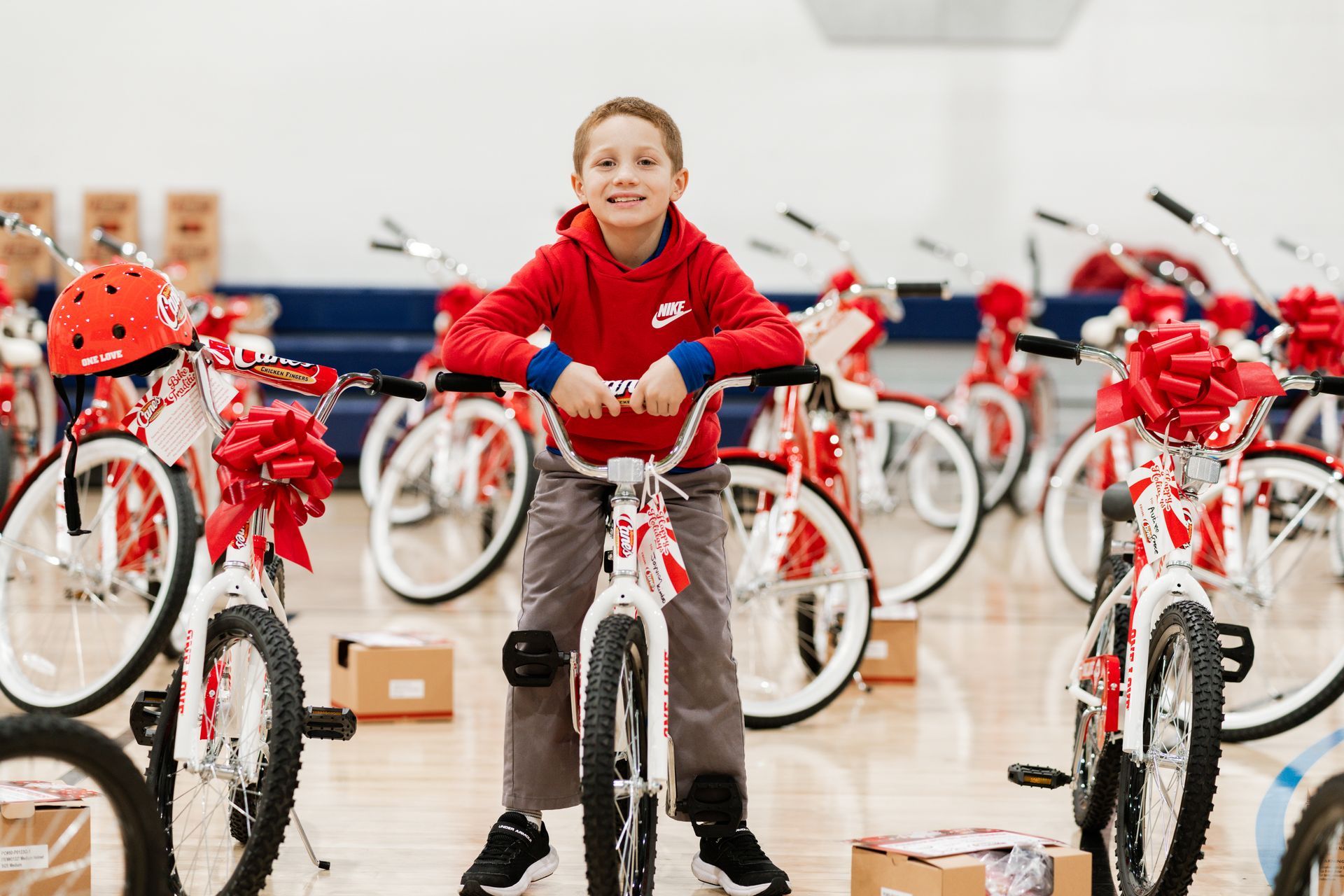 A smiling child in a red hoodie sits on a new white bicycle among rows of identical bikes in a gymnasium.