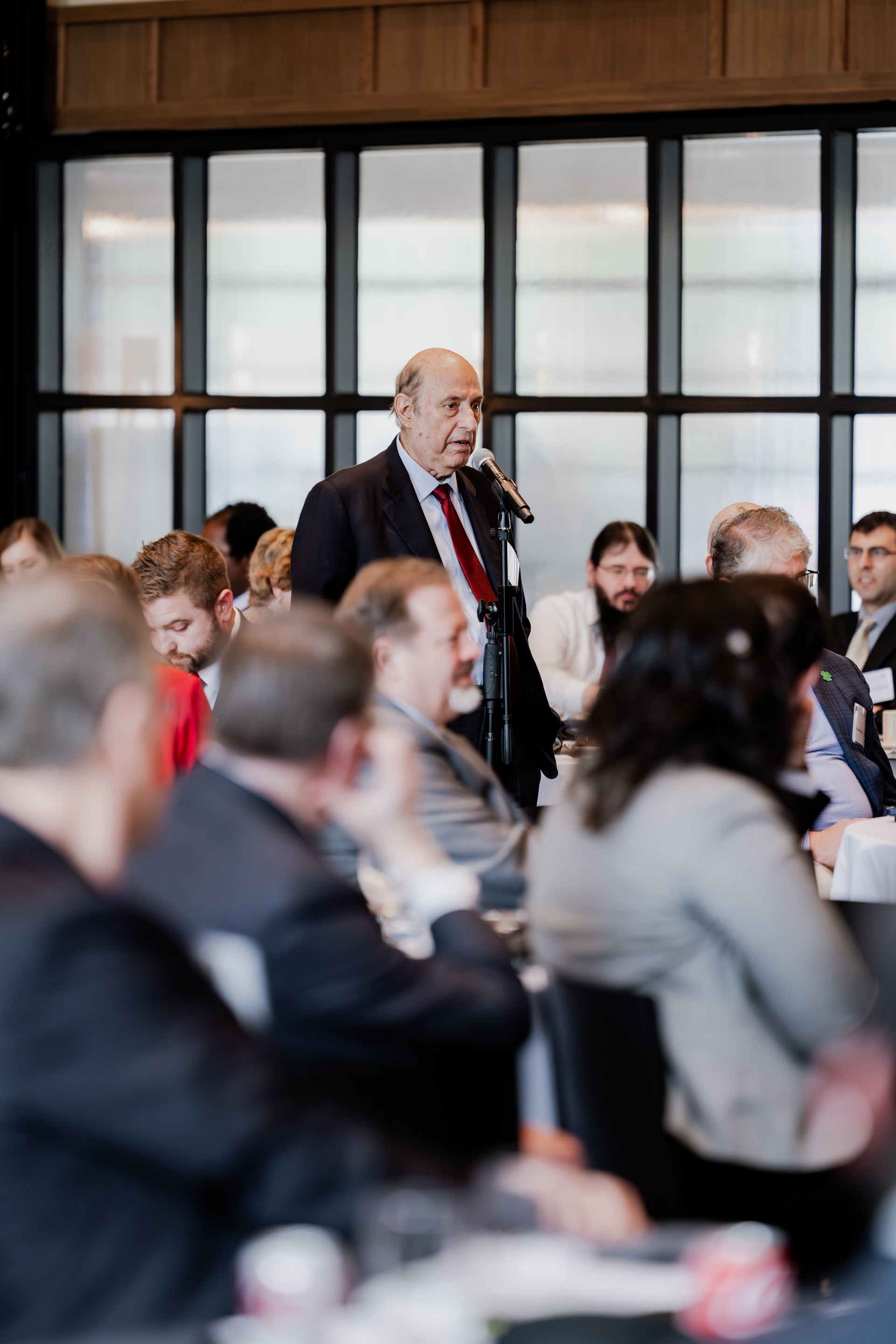 A person stands in a conference room speaking into a microphone, with a seated audience in the foreground.