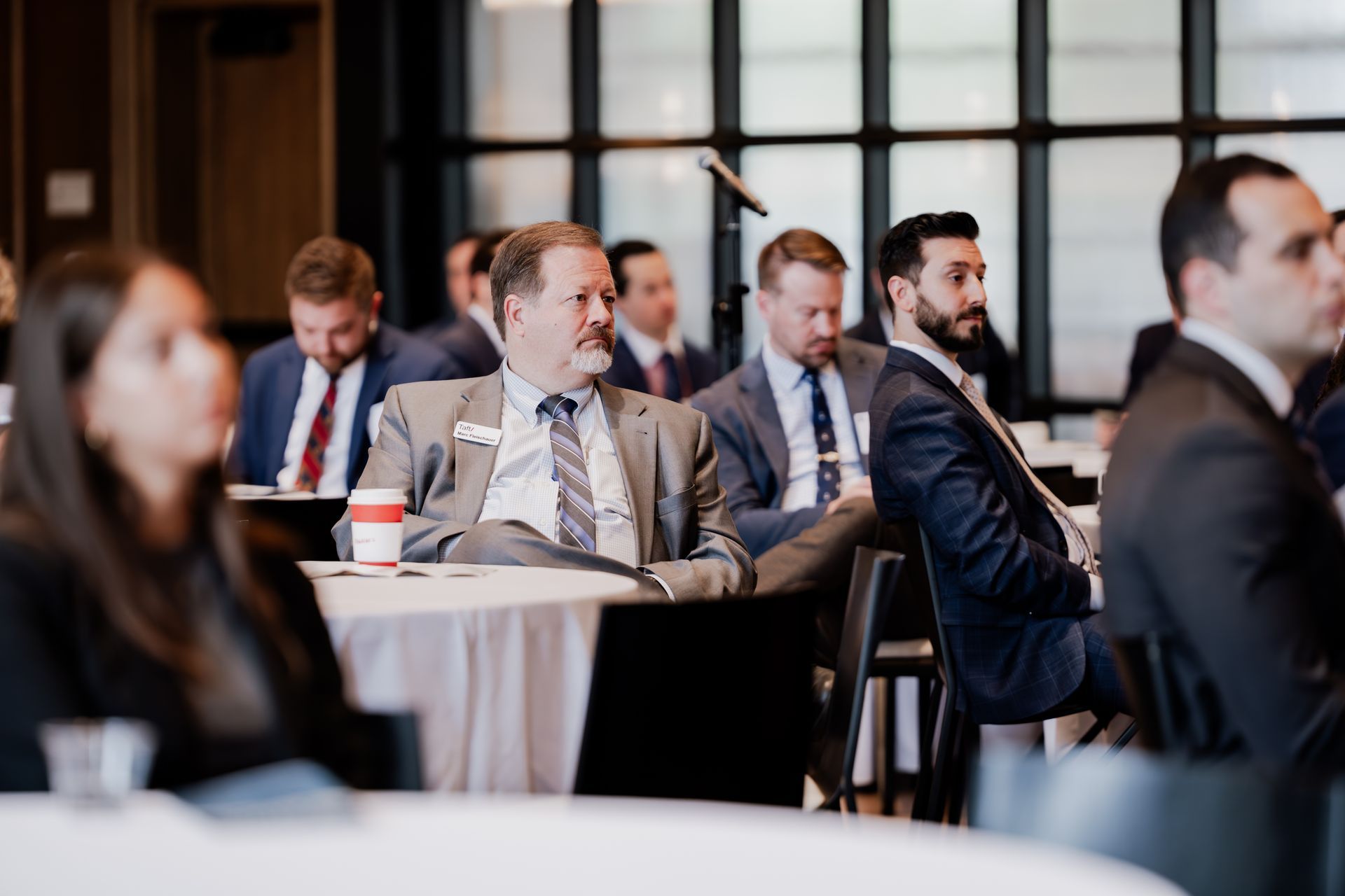 Attendees in professional business attire sit at tables during a corporate conference or seminar in a well-lit room.