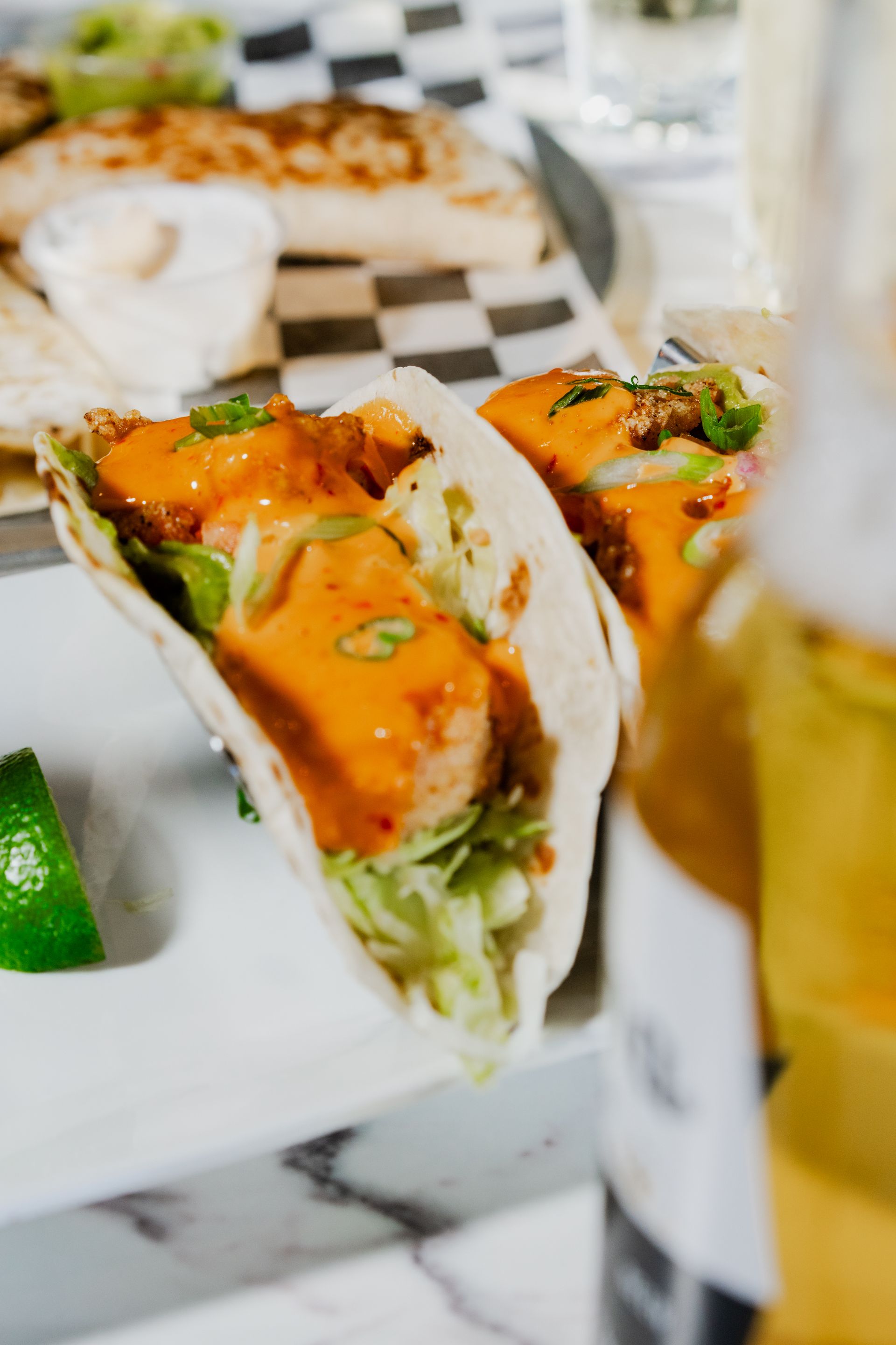 Two tacos with orange sauce on a white plate, lime wedge, beer in focus.