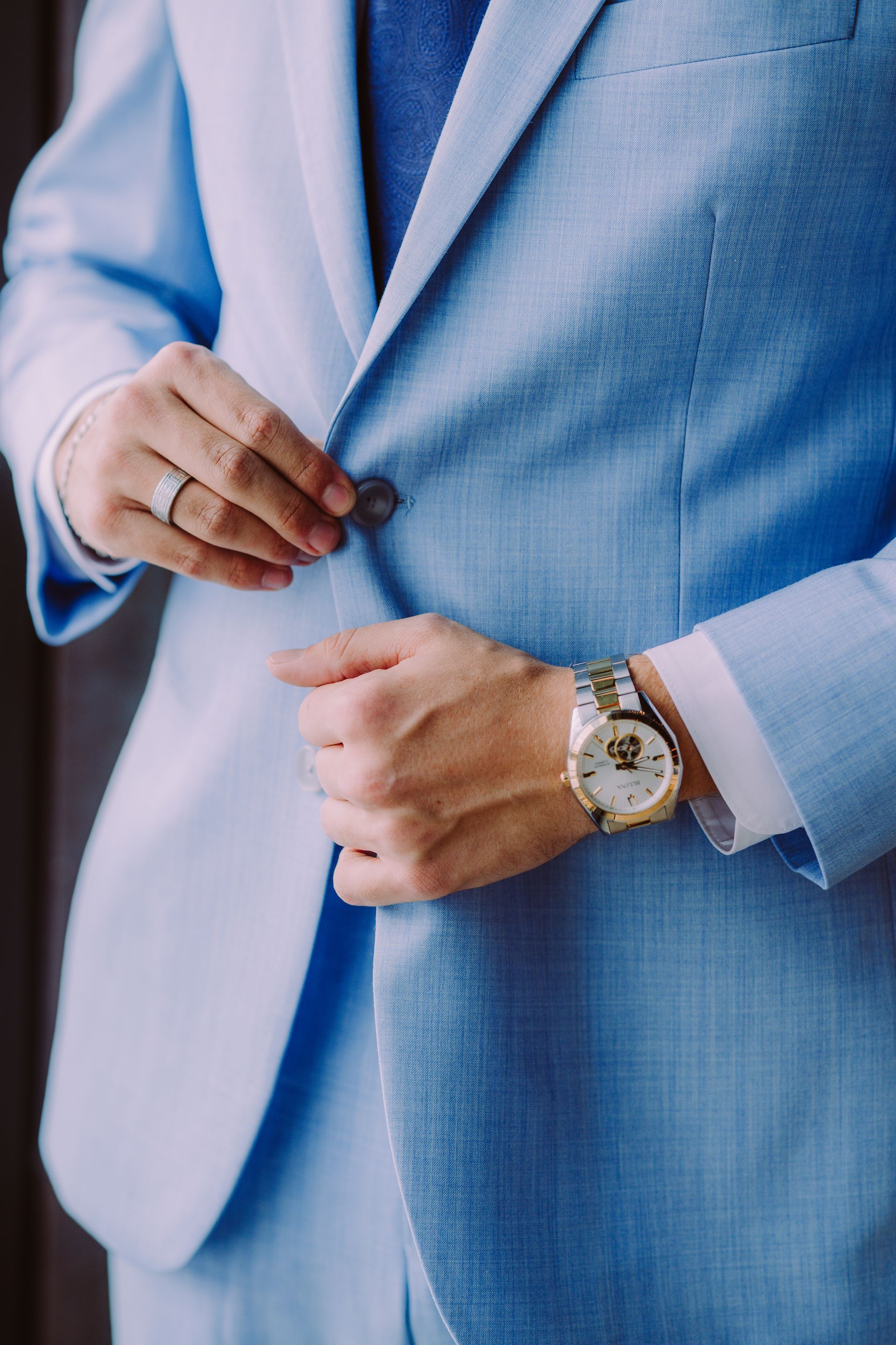 Man in a light blue suit buttoning his jacket, wearing a watch and a ring.