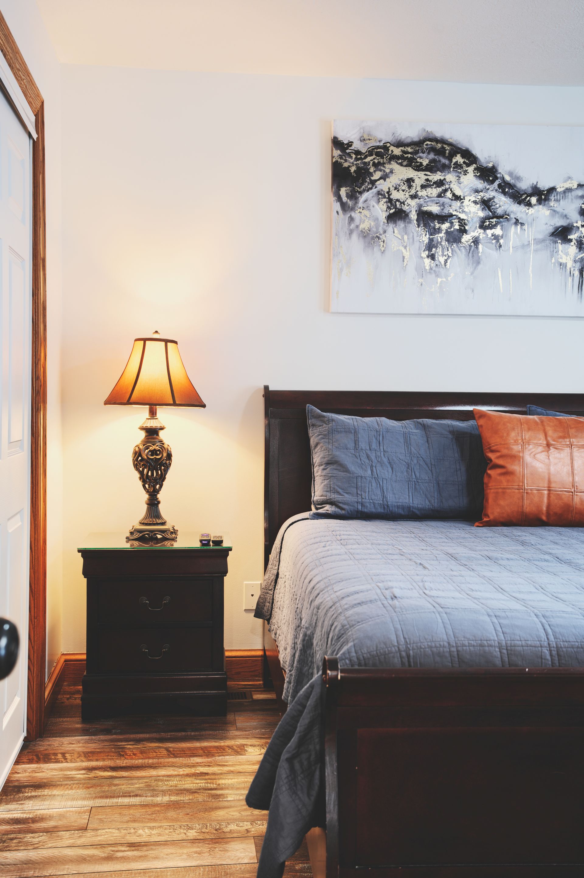 A bedroom with a bed , nightstand , lamp and painting on the wall.