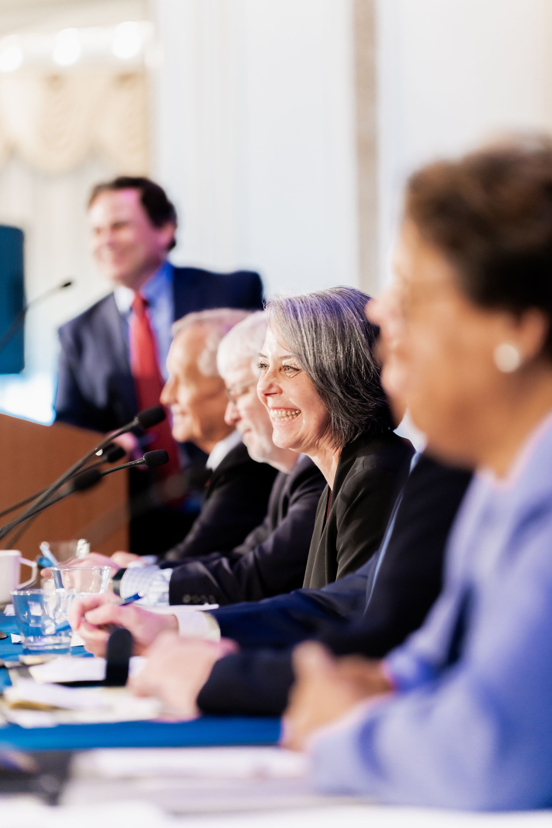 A panel of professionals sits at a table with microphones, smiling, while a speaker stands at a podium in the background.