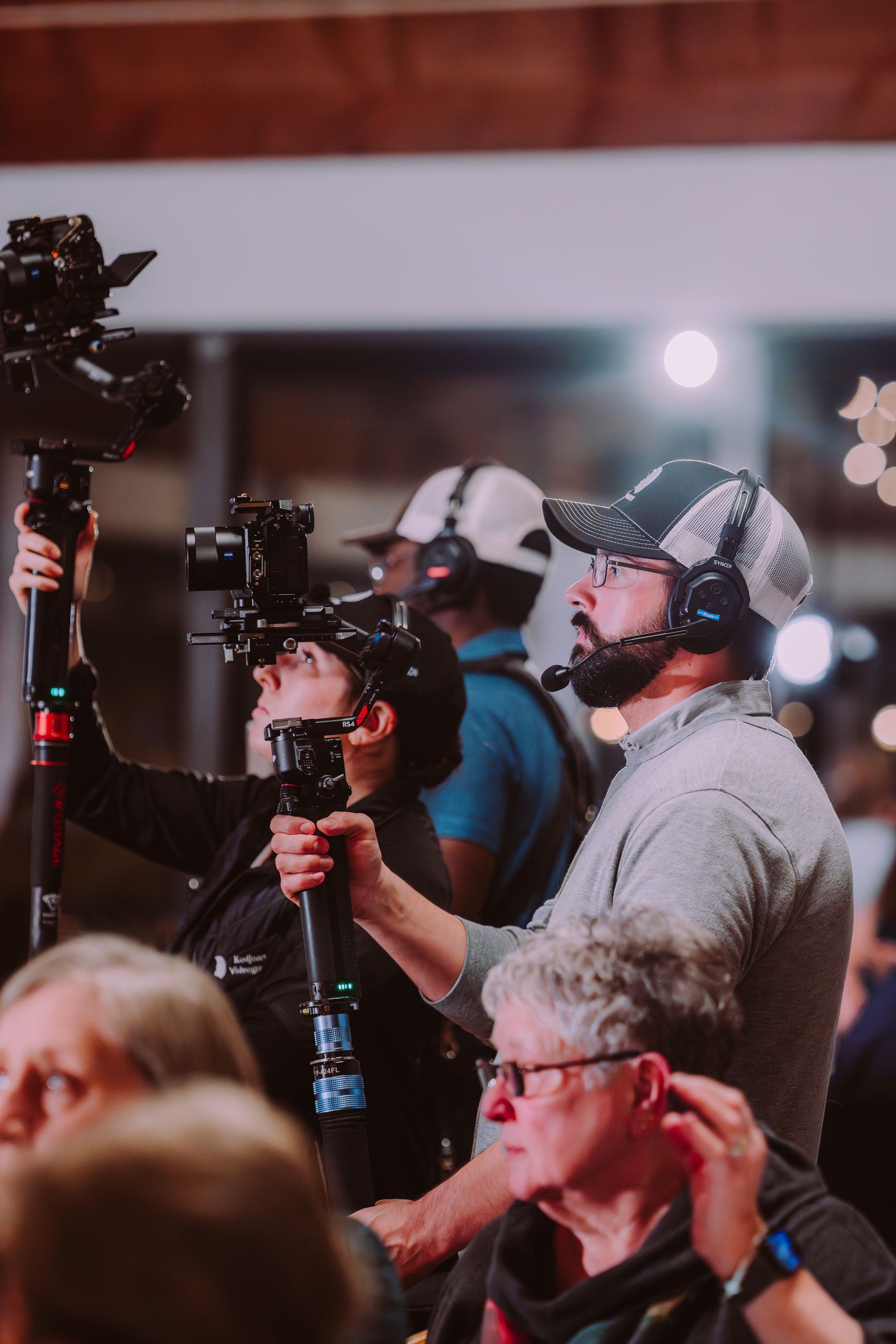 Three people hold cameras on poles, filming an event. One man wears a hat and headset. Blurred background.