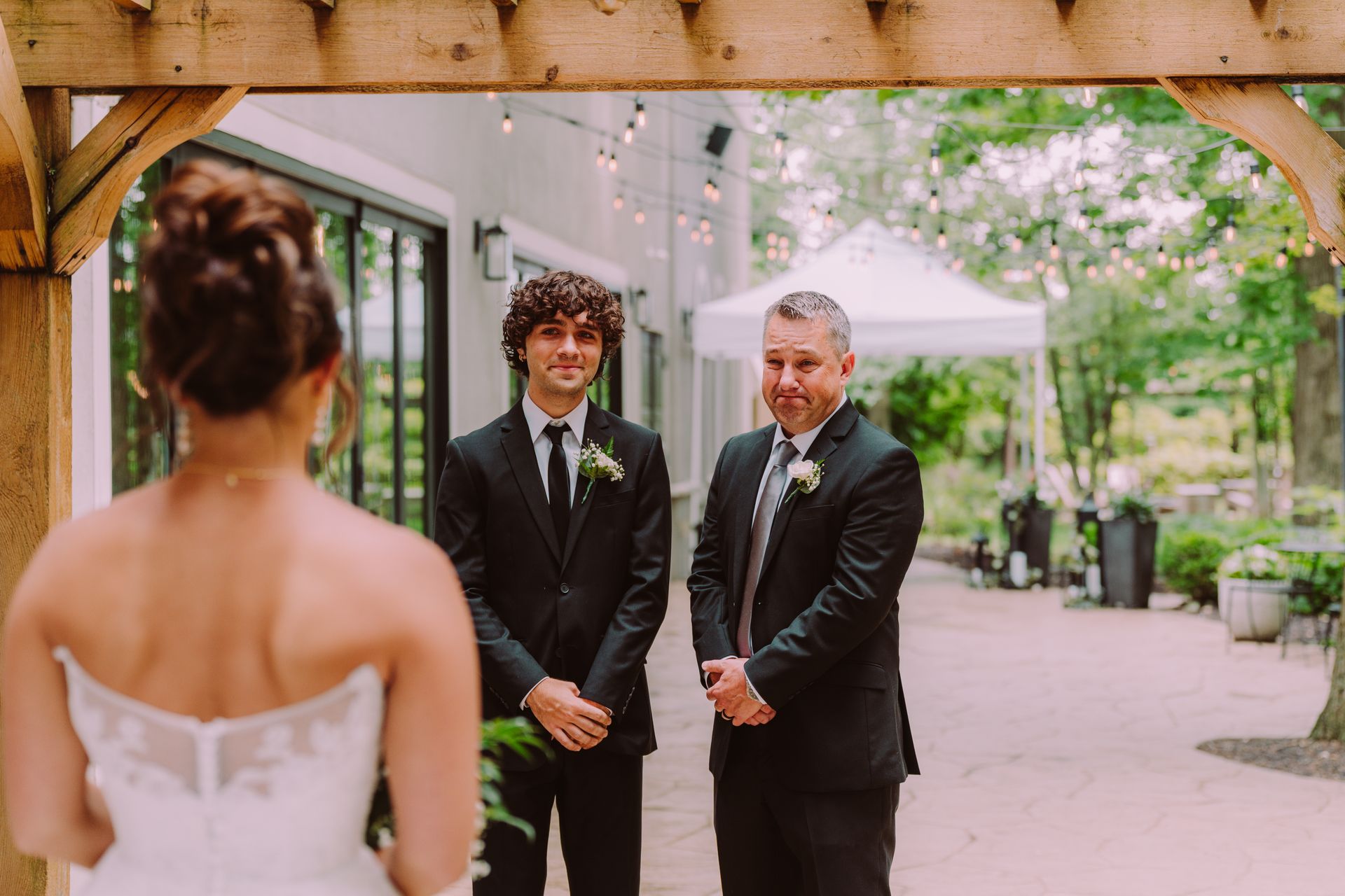 Bride facing the groom and father under an arbor. Groom and father in suits, outdoor setting.