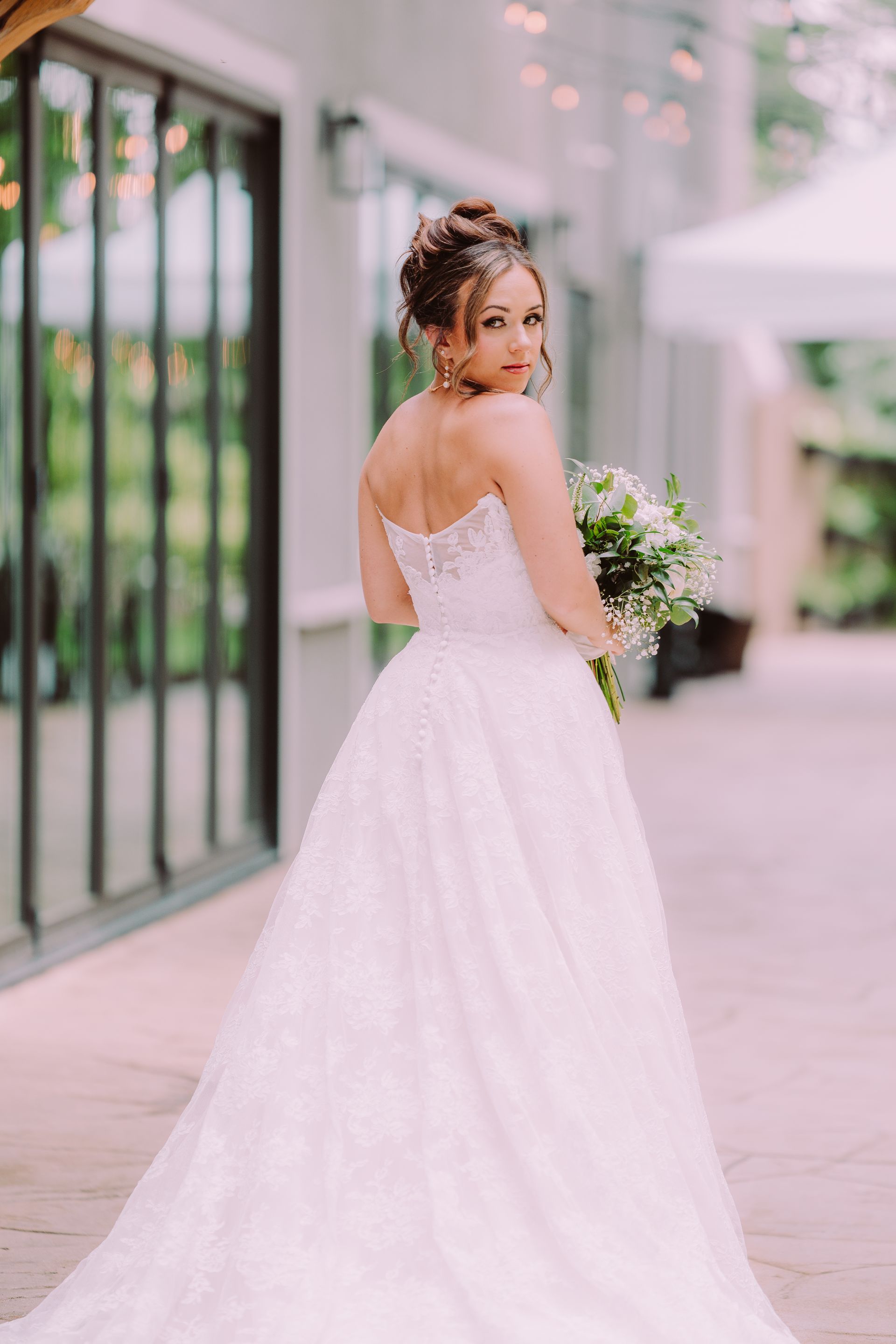 Bride in a white strapless wedding dress, holding a bouquet, looks back over her shoulder with a gentle smile.