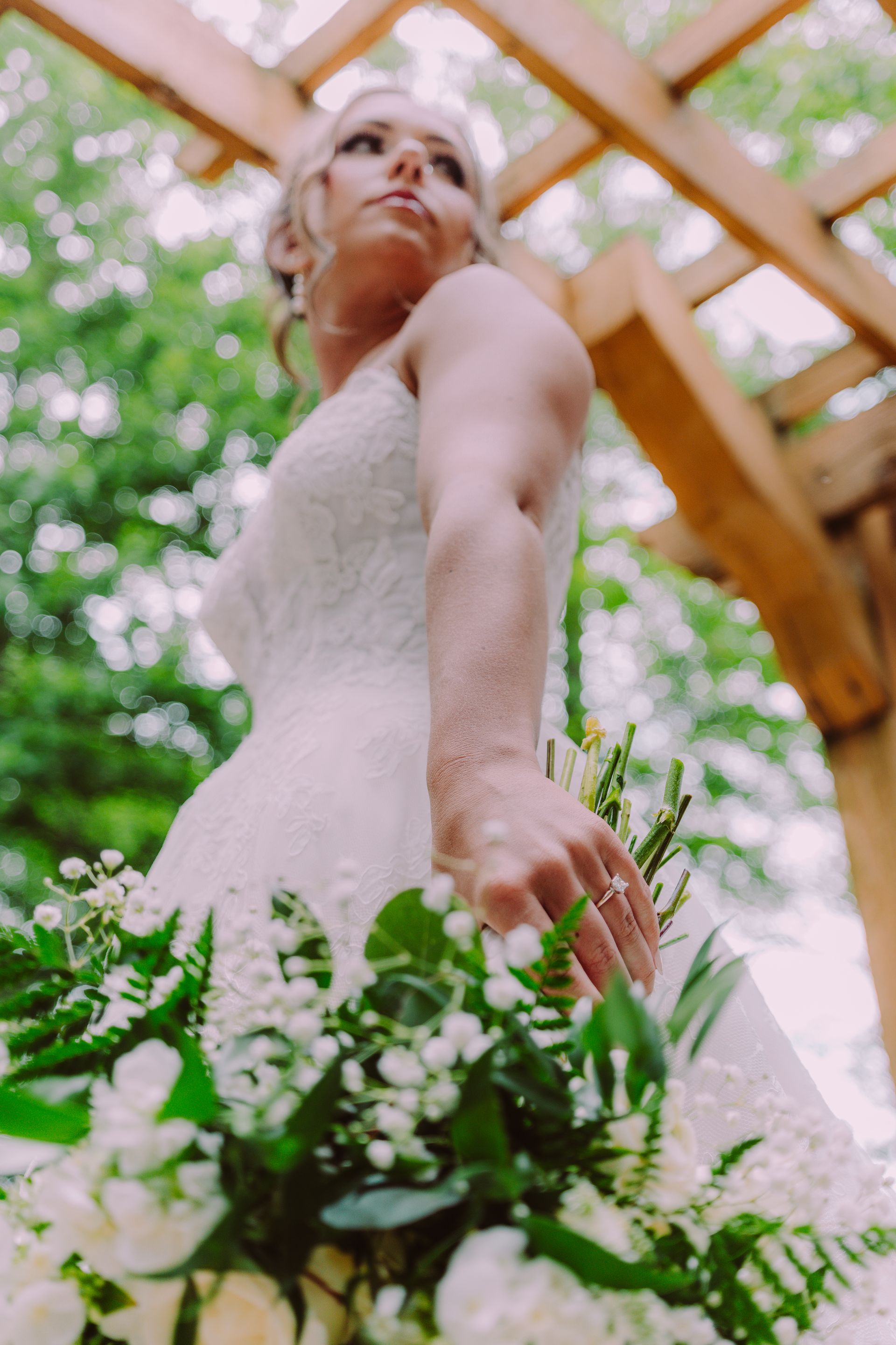 Bride in a white wedding dress holds a bouquet, looking upwards in a garden setting.
