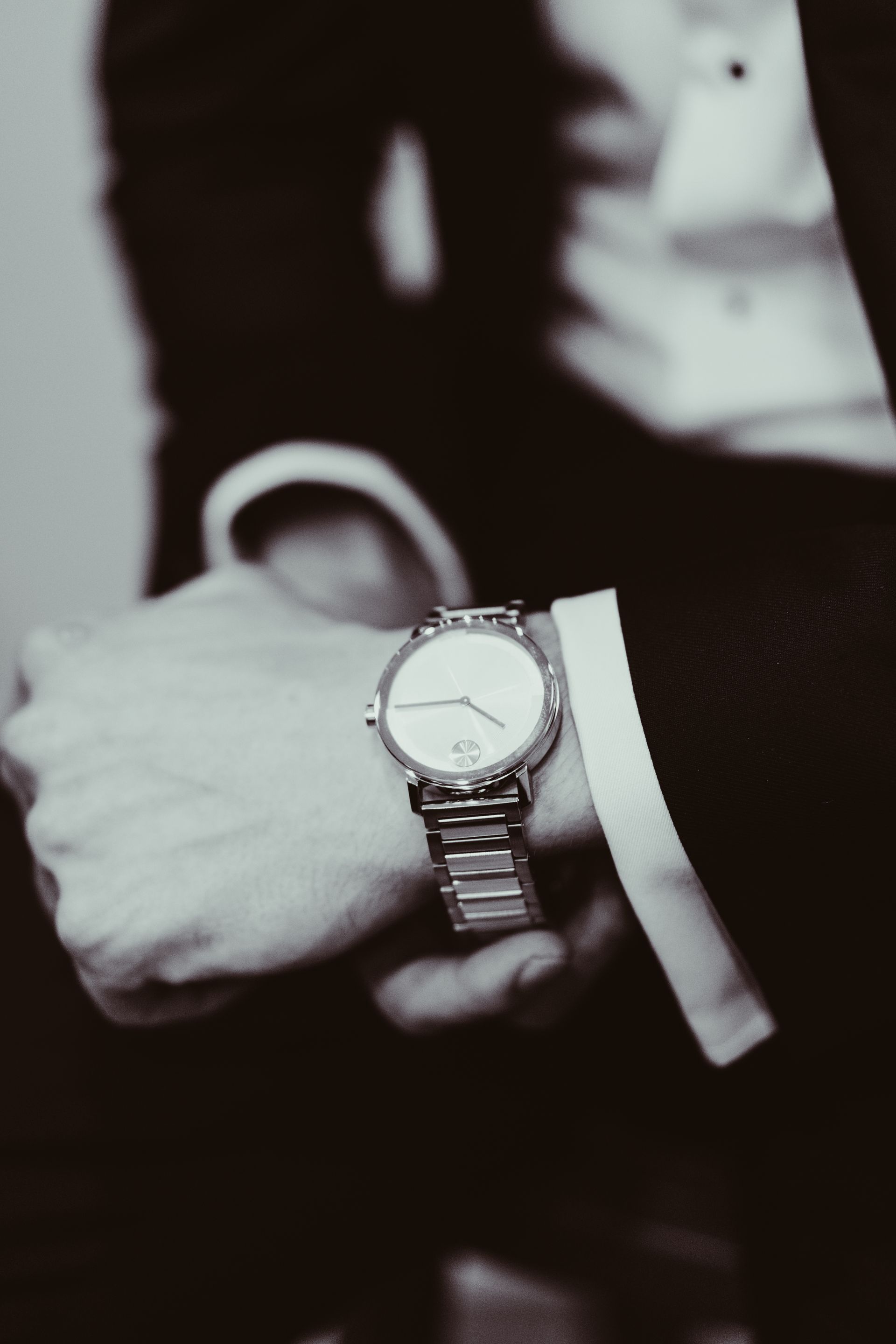 Close-up shot of a man's wrist with a silver wristwatch, part of a formal suit. Black and white.