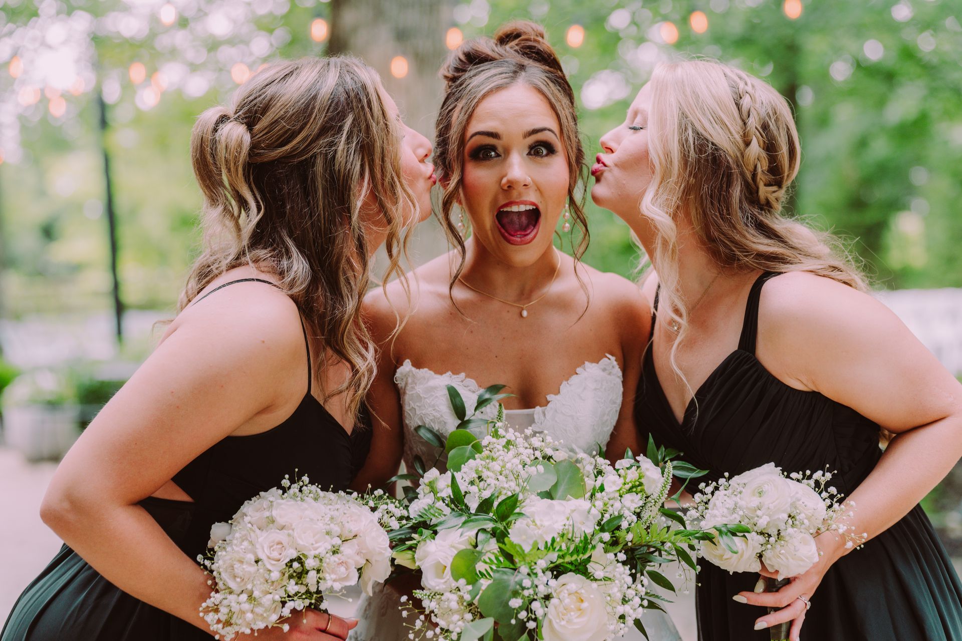 Bride with open mouth and two bridesmaids kissing her cheeks; holding white floral bouquets; outdoor setting.
