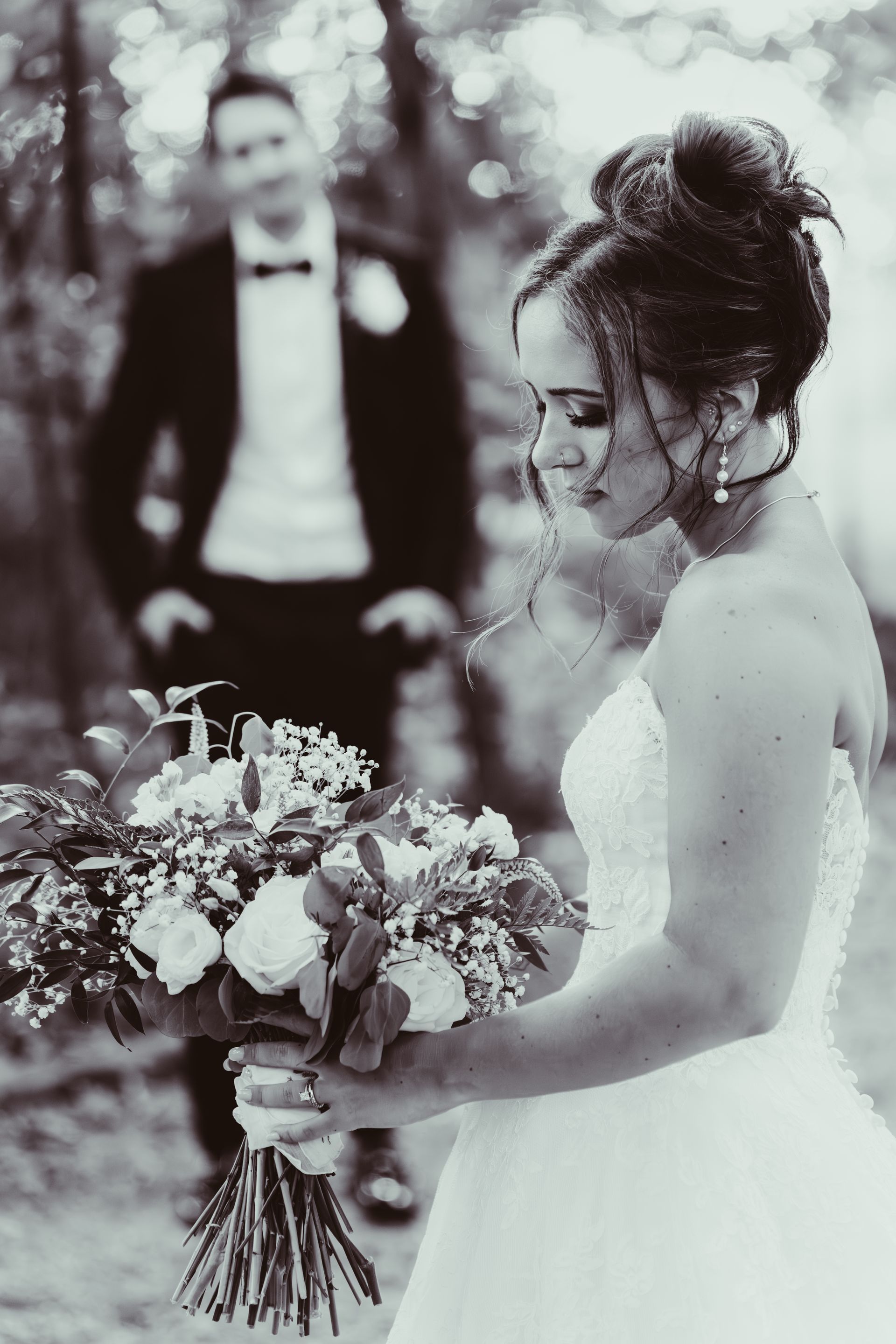 Bride in a white gown holding a bouquet, looking down. Groom in a tuxedo, standing behind her. Black and white photo.
