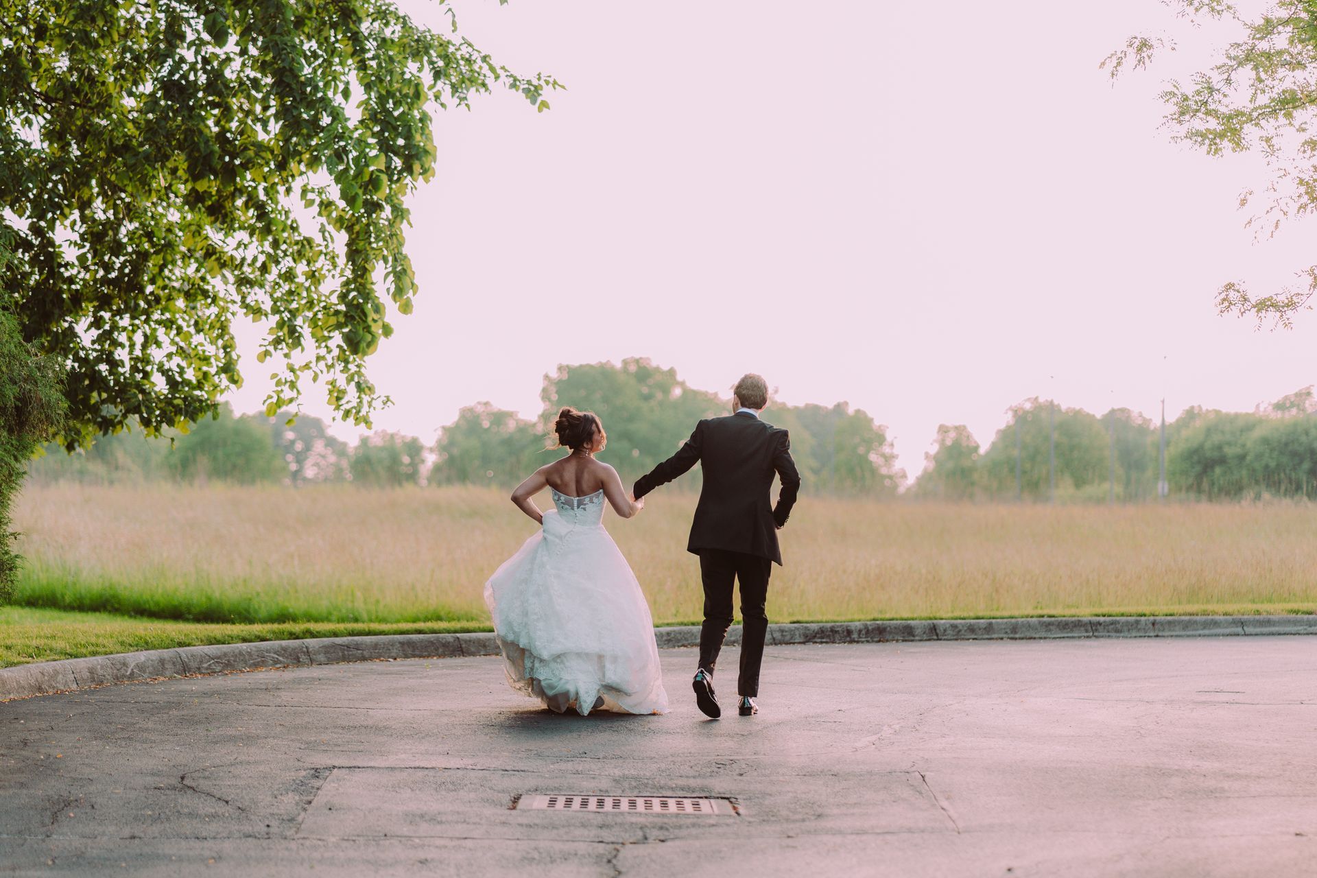 Newlyweds run hand-in-hand across a paved area towards a field. 