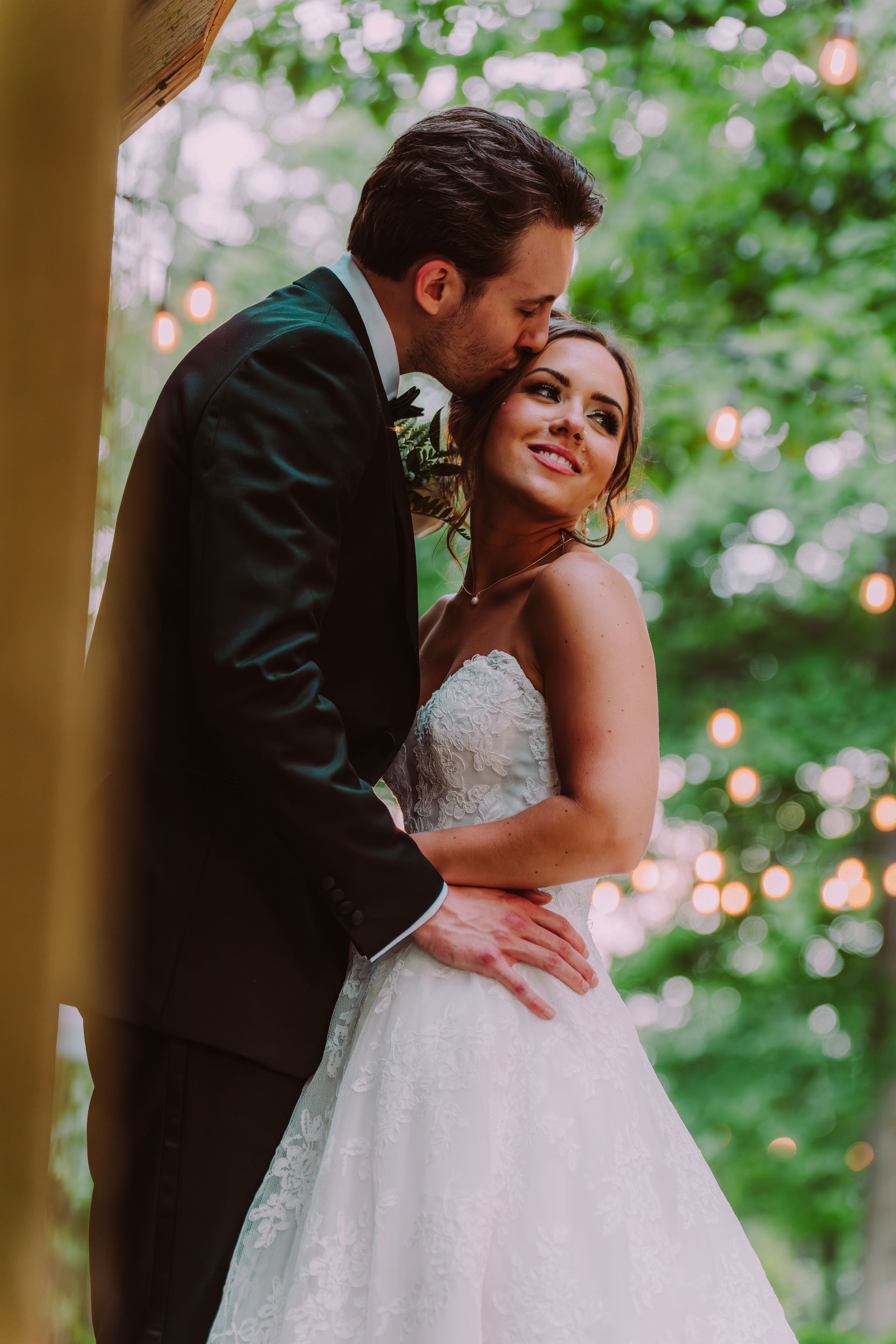 A groom in a black tuxedo kisses the forehead of his bride, who wears a white beaded gown.