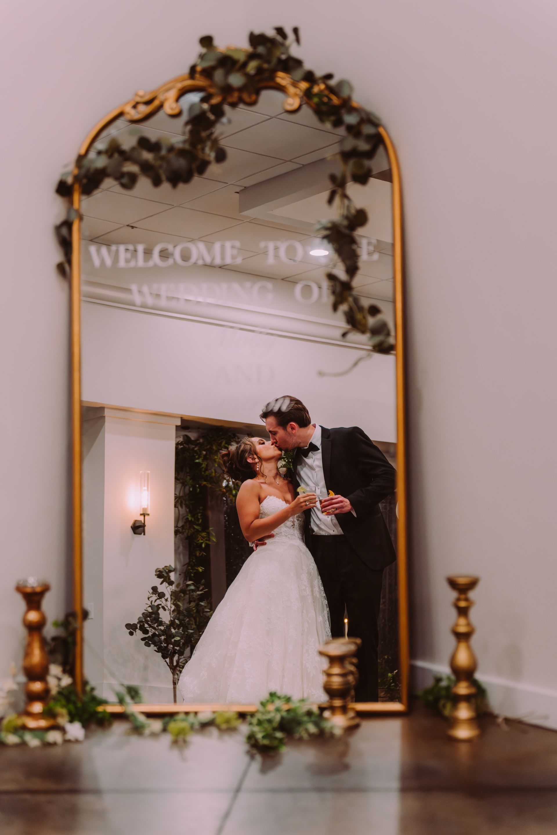 Bride and groom kissing reflected in a gold-framed mirror. The mirror reads 