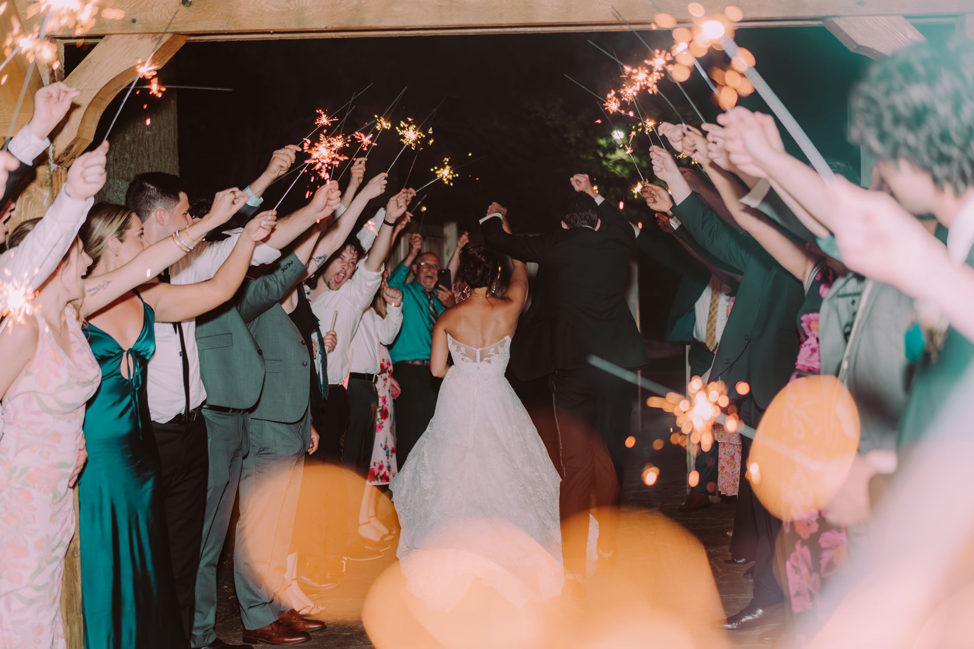 Bride and groom exiting wedding reception through a sparkler tunnel, guests cheering.