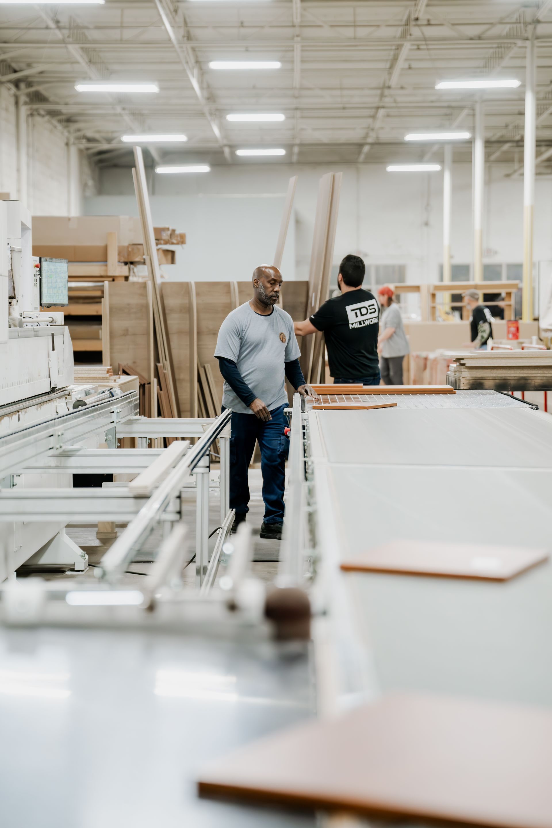 Man in grey shirt standing near industrial machinery in a woodshop. Others work in the background.