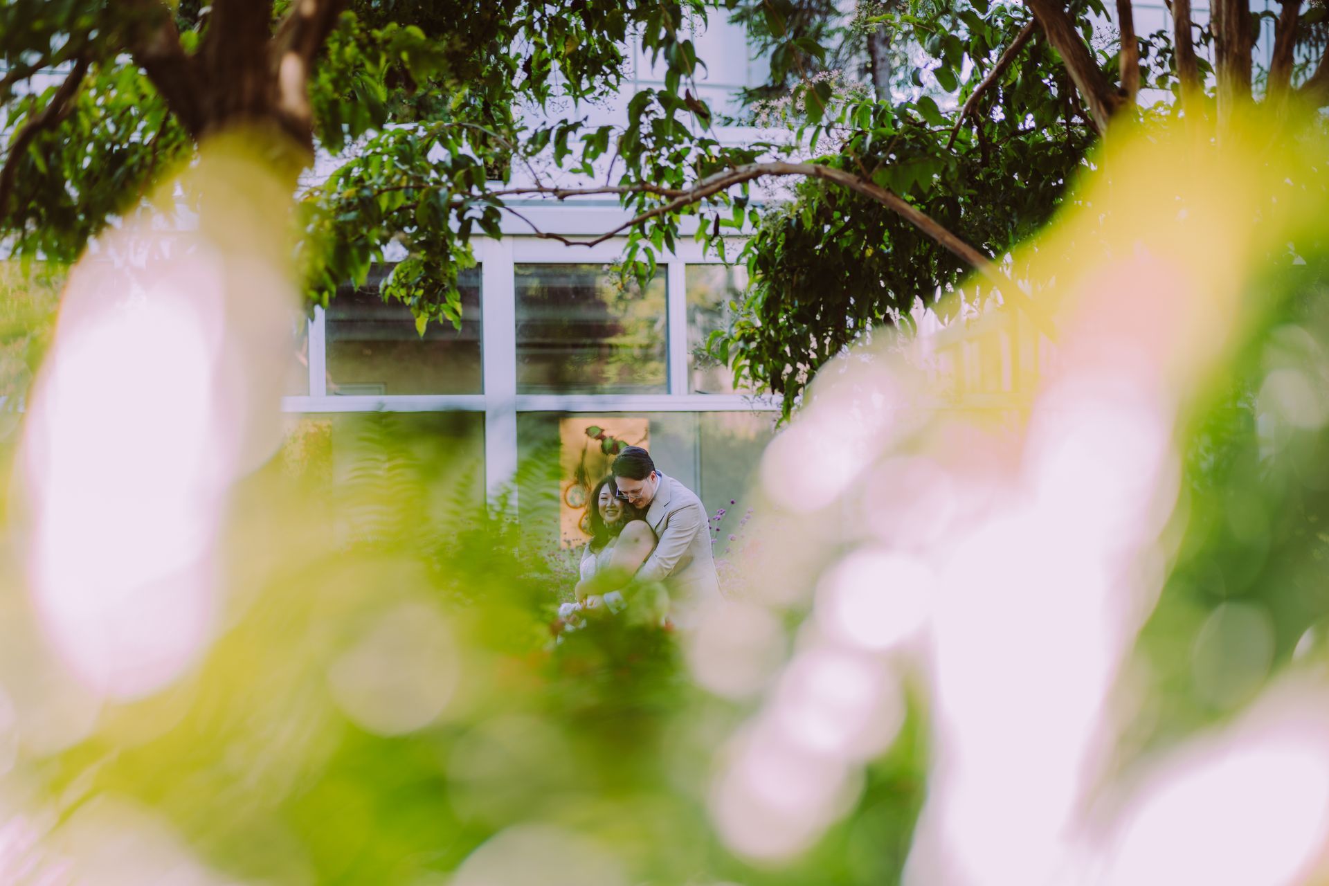 Couple hugging by a window framed by greenery. Bright, sunny outdoors; yellow and green hues.