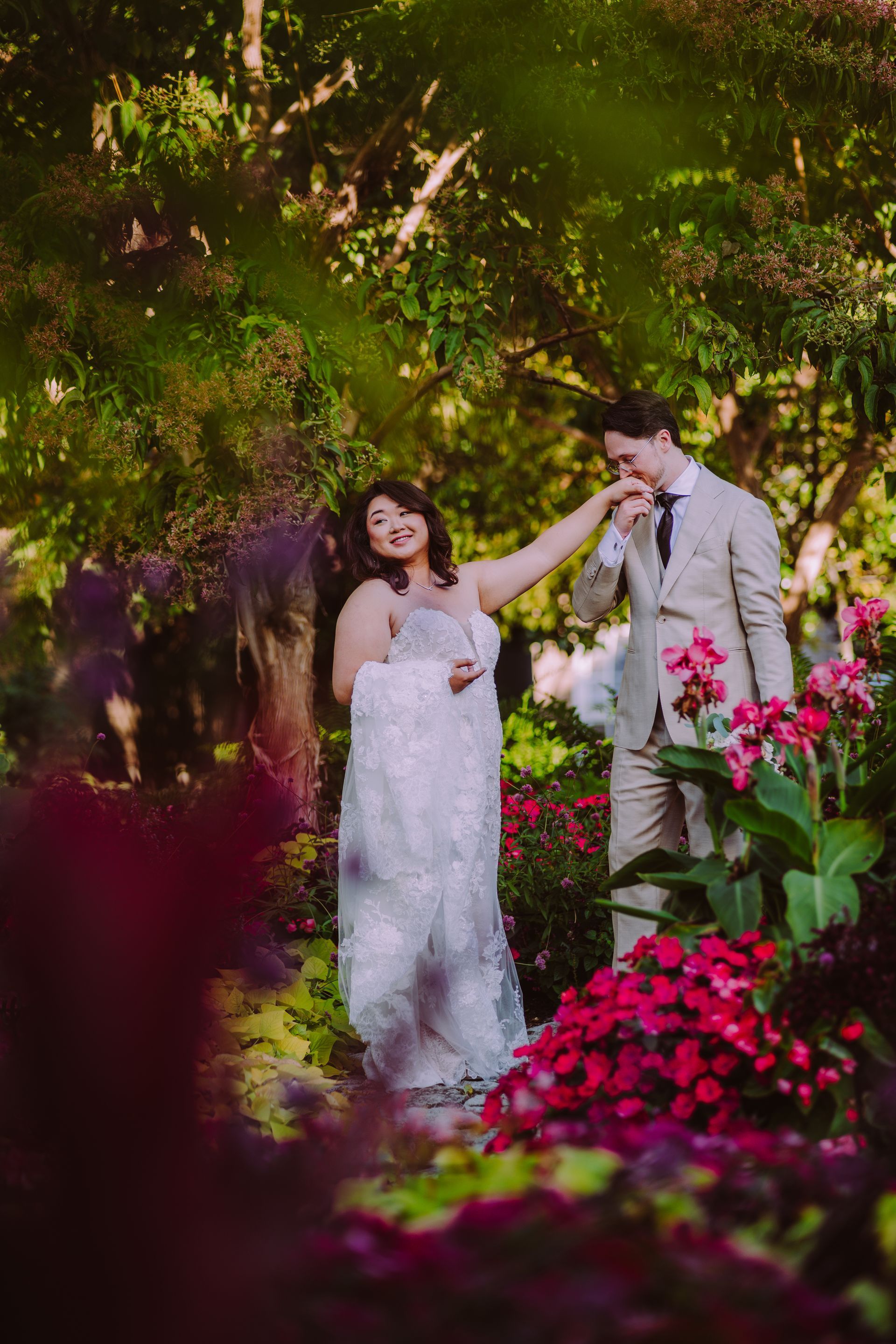 Couple in formal attire, woman in white dress, man in tan suit, pose in garden with red flowers.