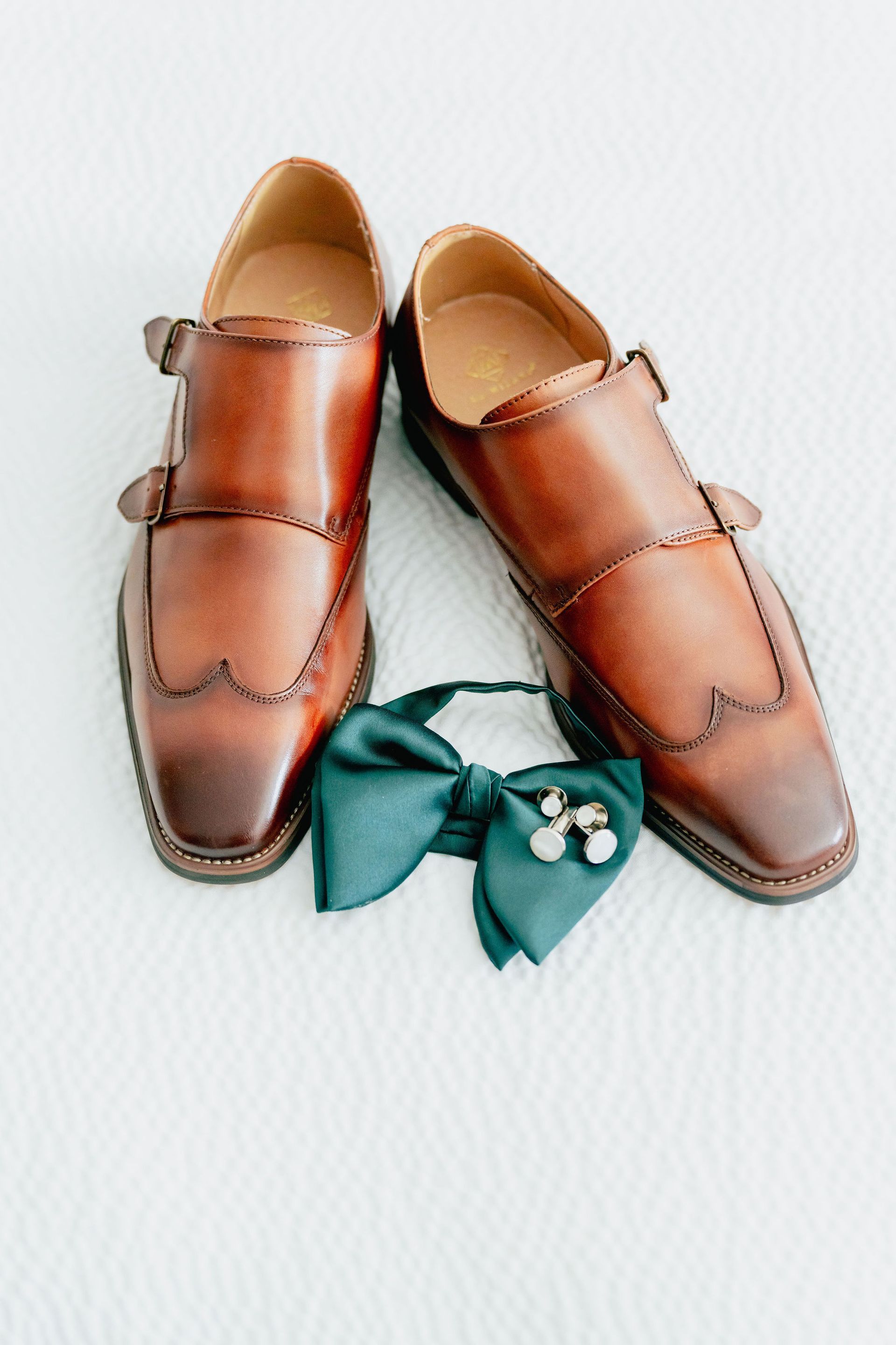 A pair of brown shoes and a green bow tie are sitting on a bed.