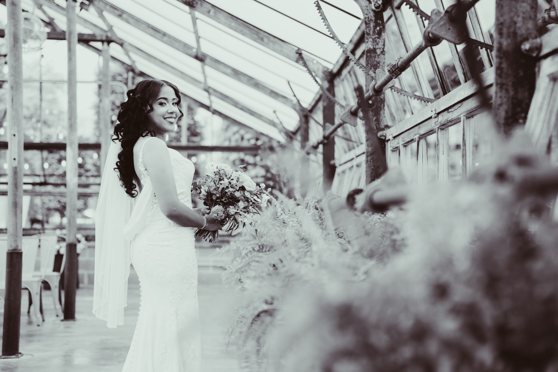 Bride in white dress smiles, holding flowers, inside a greenhouse.