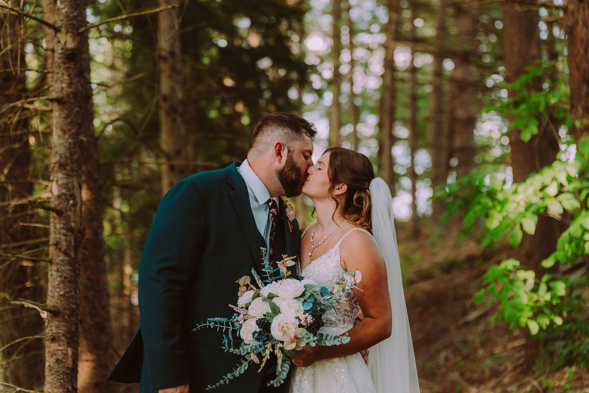 Couple kissing amidst a forest. The groom wears a dark suit, the bride a white dress and veil, holding a bouquet.