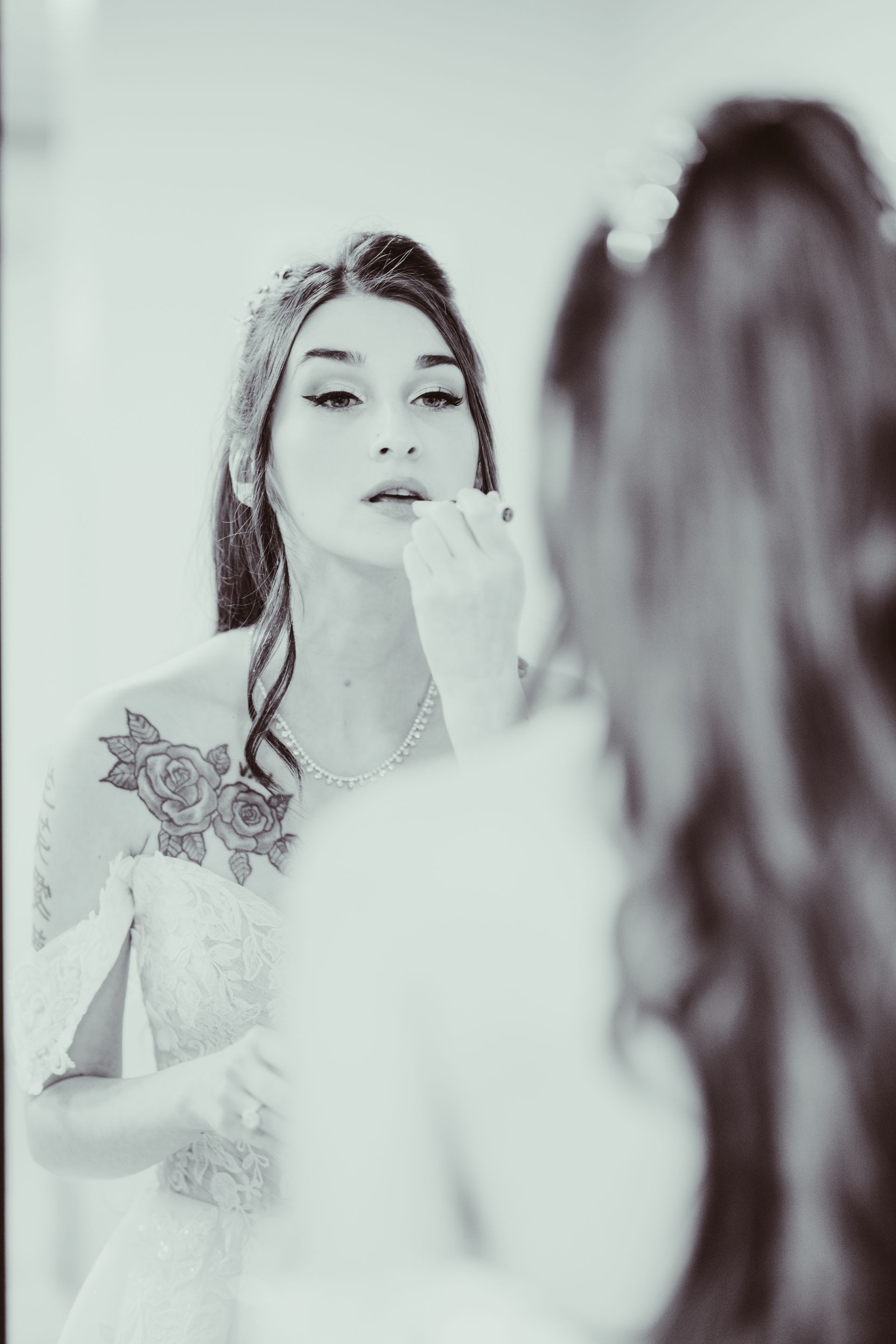 Woman applying lipstick in a mirror; floral tattoo on her shoulder, formal gown.