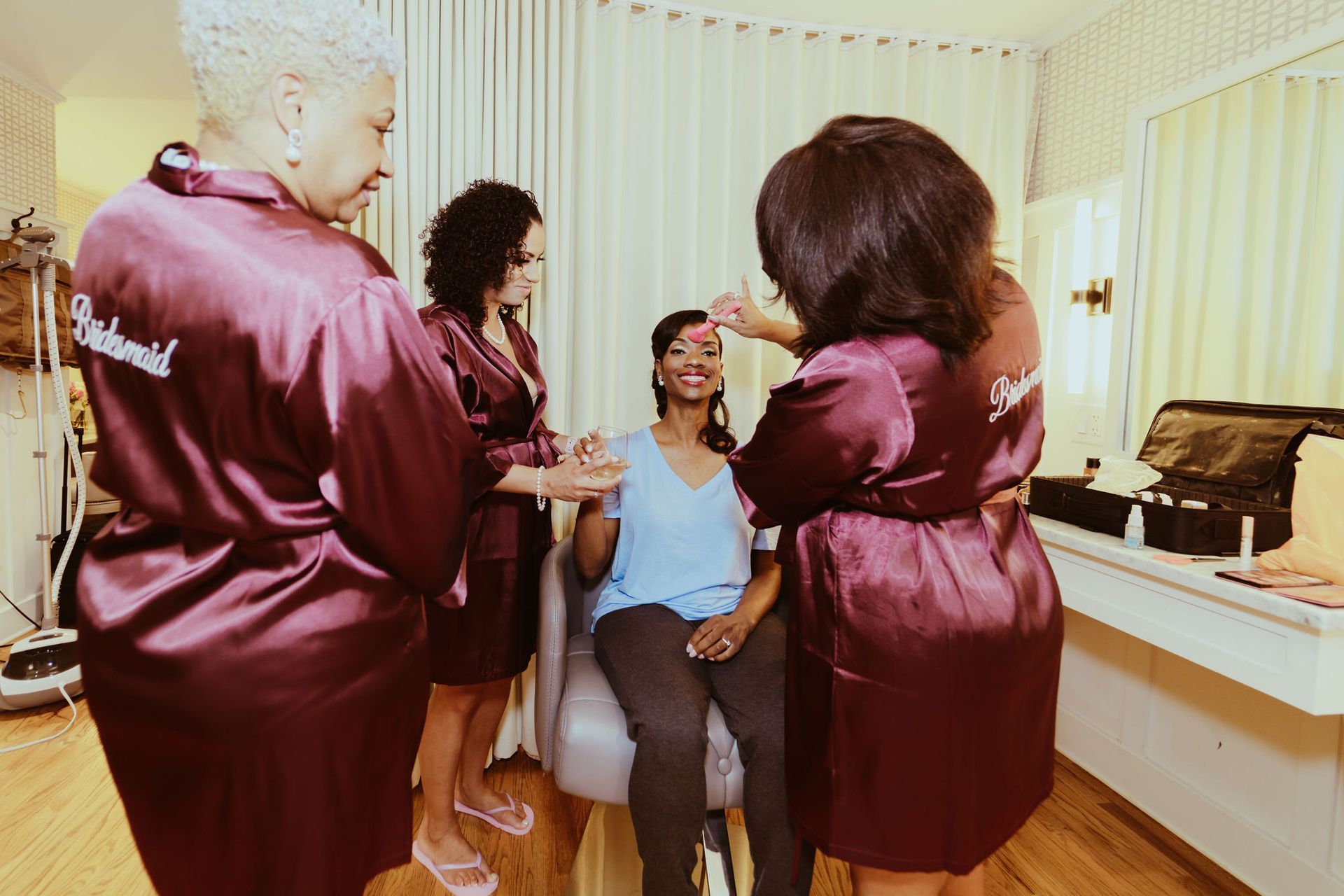 A group of women are standing around a woman sitting in a chair getting her makeup done.