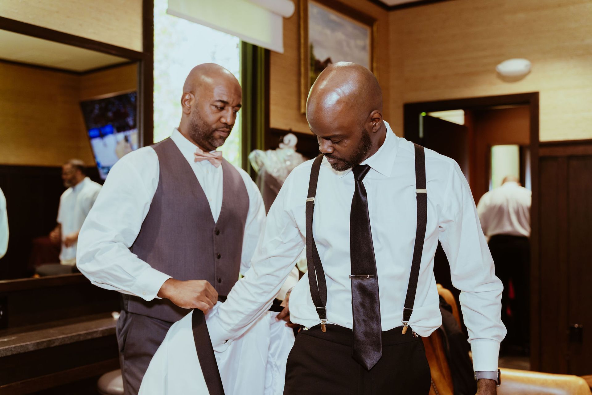 Two men in suits and suspenders are getting ready for a wedding.