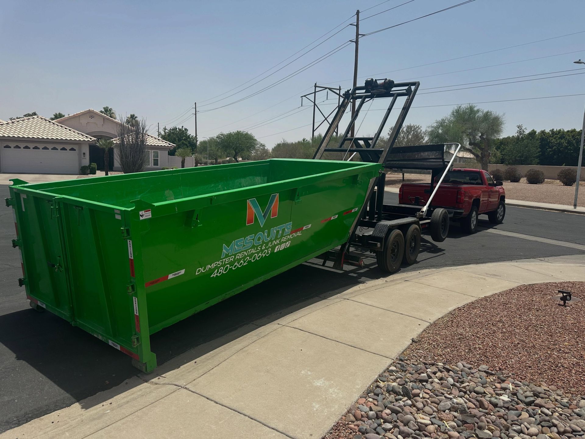 Green dumpster being hauled by a red tow truck on a suburban street.