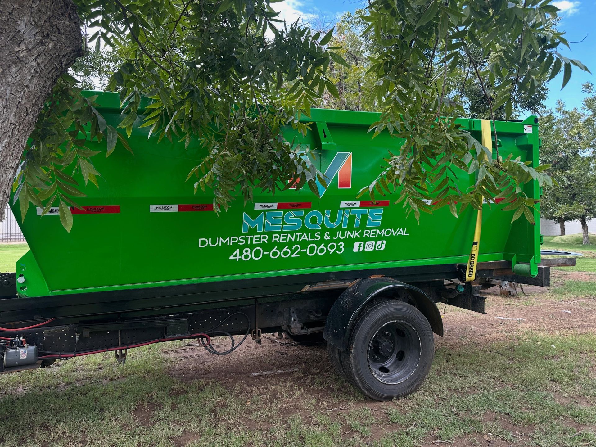 Green dump trailer parked under trees with business lettering on the side