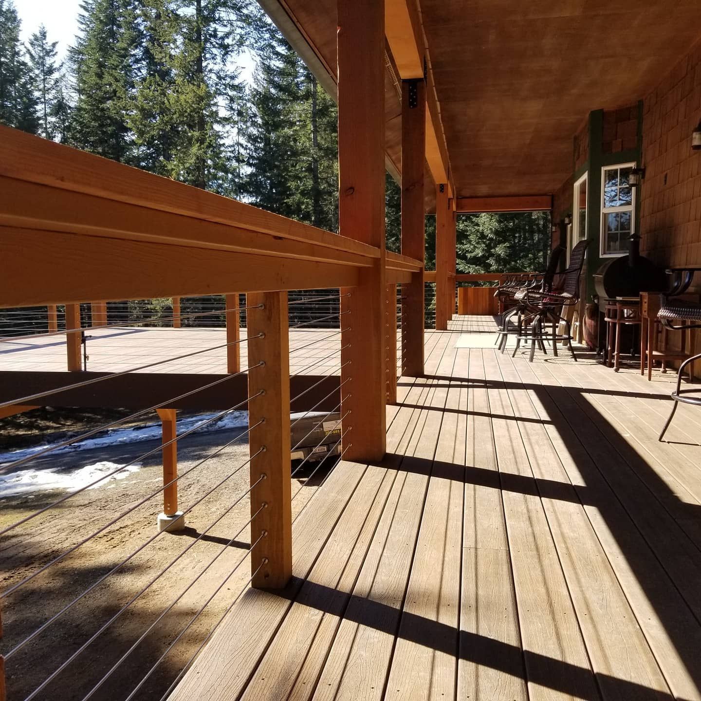 Wooden deck with cable railing and roof. Chairs and table visible on the deck's far end, with a forest in the background.