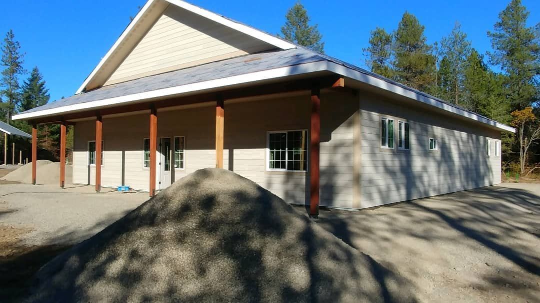 Building with a peaked roof and covered porch. Tan walls, brown posts, blue sky. Gravel pile in foreground.