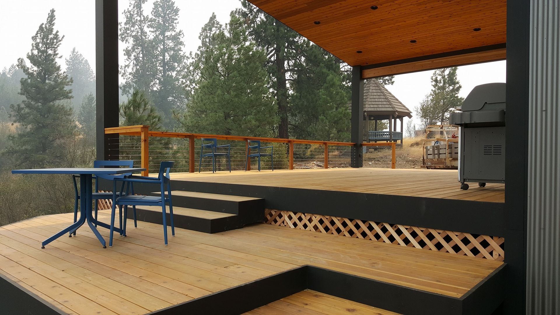 Wooden deck with blue table/chairs, leading to a covered patio. Grill, trees in background.