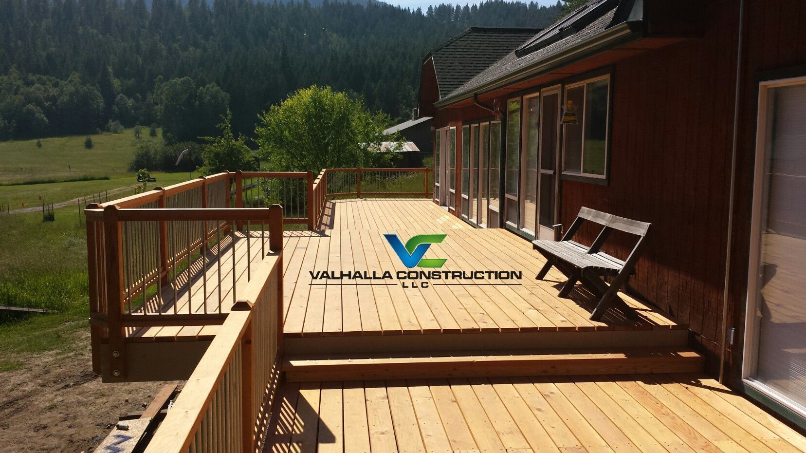 Wooden deck with railing attached to a reddish-brown house, bench, and scenic backdrop.