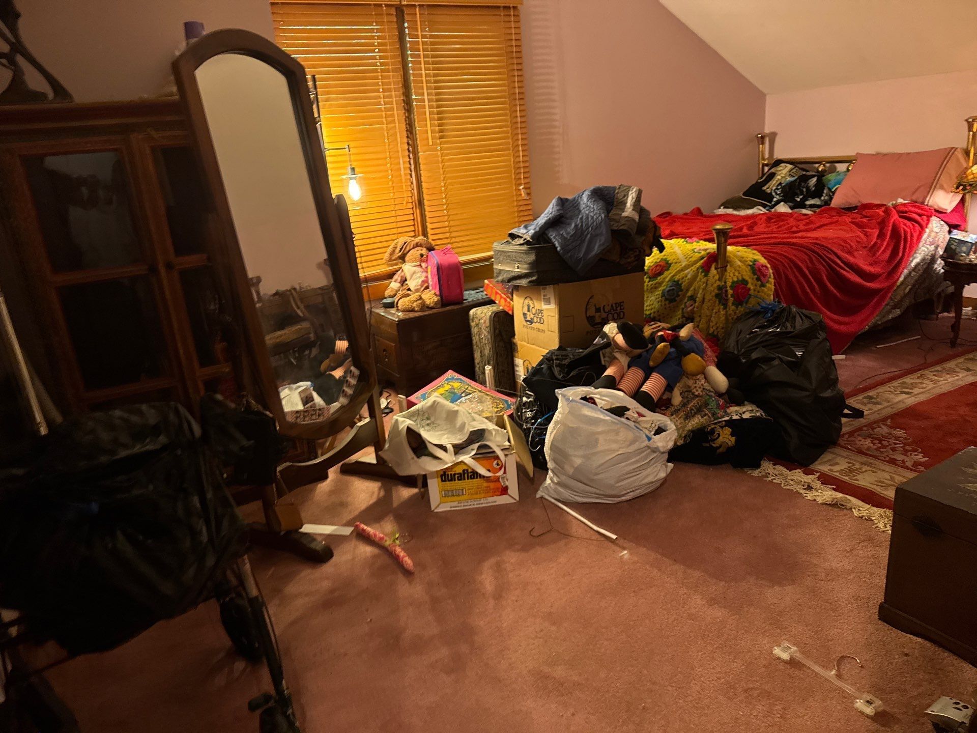 Cluttered bedroom with a bed, mirror, chair, and piles of clothes and bags on the floor