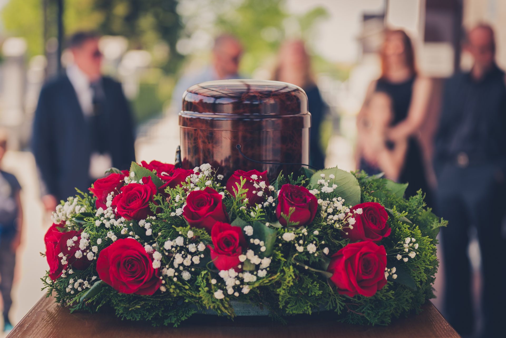 Flowers arranged around a polished urn at an outdoor memorial service with blurred mourners in the background