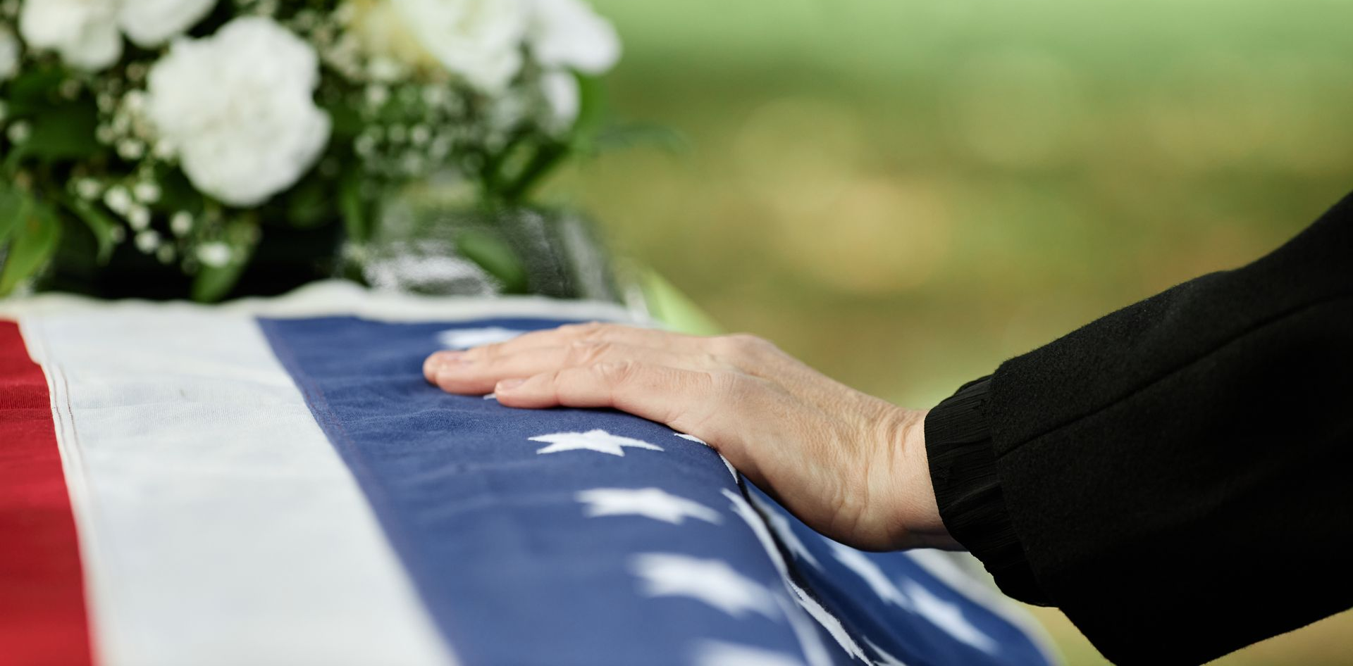 Hand resting on a coffin draped with a red, white, and blue flag at a funeral